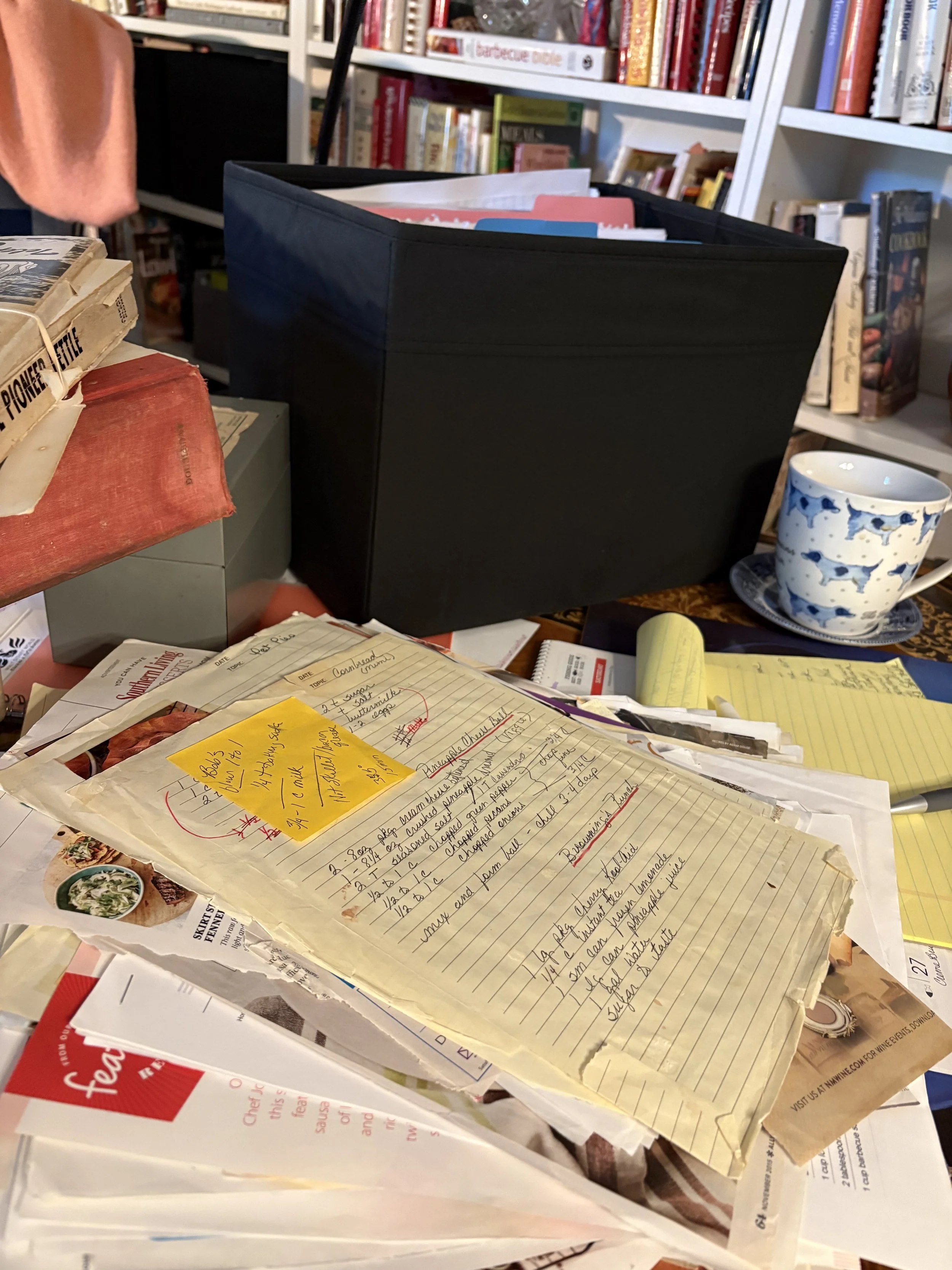 A well-used recipe workspace with handwritten recipe cards, vintage cookbooks, a box of family recipes, notebooks and a coffee cup, capturing  the work behind preserving generations of family recipes.
