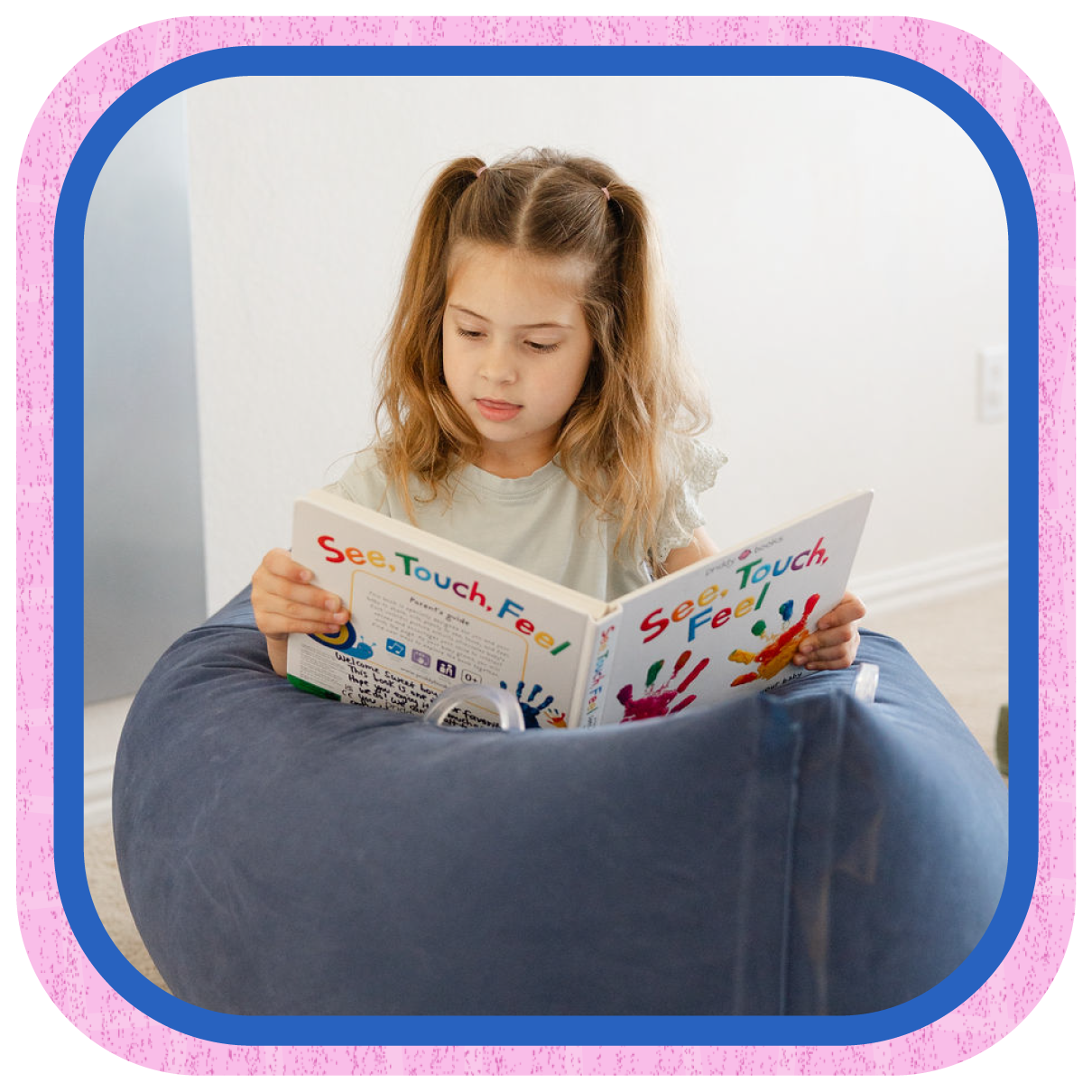 A young girl with light brown hair in pigtails reading a colorful children's book titled 'See, Touch, Feel' while sitting on a large bean bag chair in a bright room.