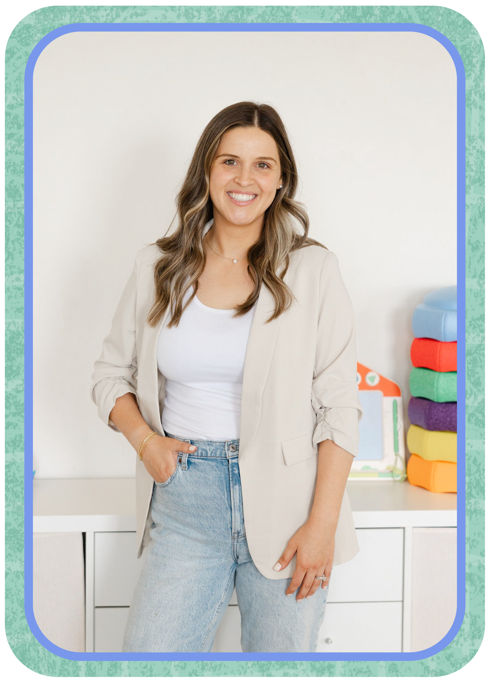 A woman with light brown hair smiling and wearing a beige blazer over a white top, with light blue jeans, standing in front of a colorful set of stacked fabric cubes and a white cabinet or drawers.