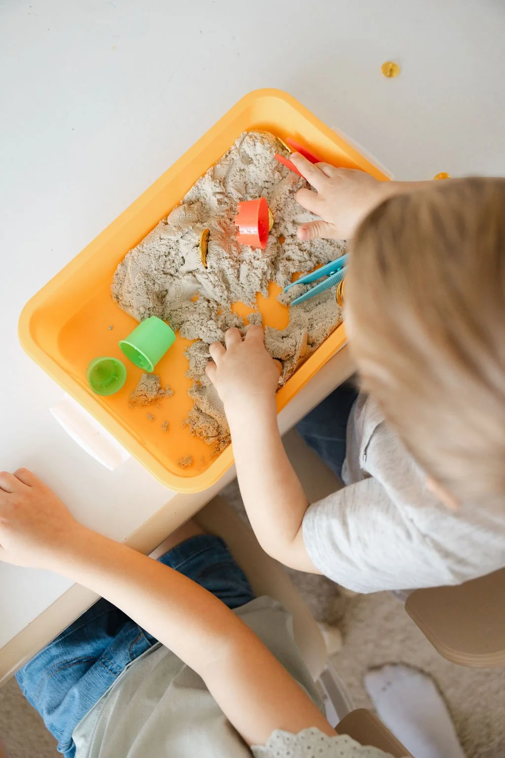 Child playing with sand in an orange sandbox with colorful plastic toys.
