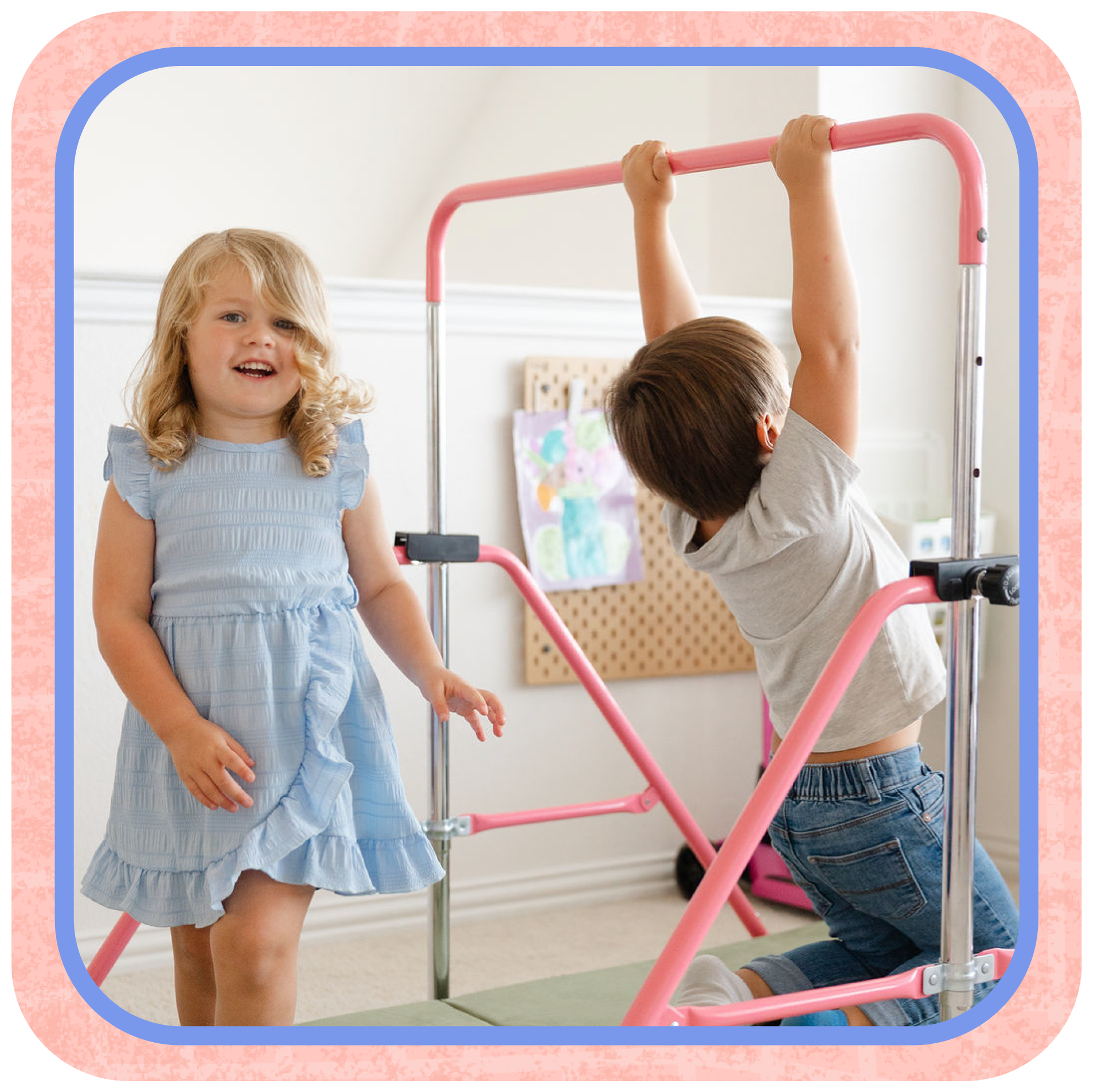 Two children playing on a pink indoor climbing frame. A girl with blonde hair in a blue dress stands and smiles, a boy with brown hair in a grey shirt and jeans is climbing and gripping the top bar.