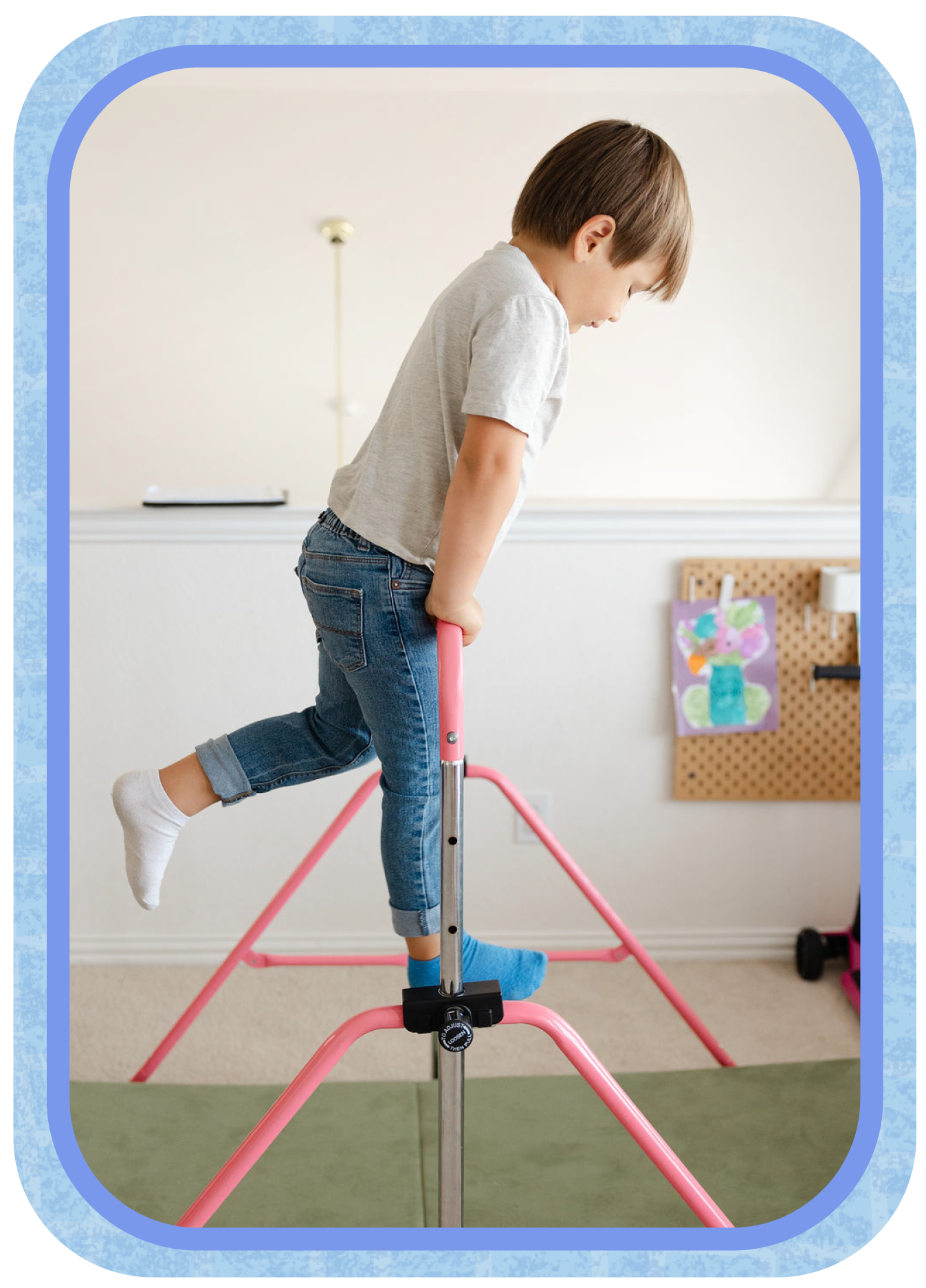 A young boy is practicing on a pink and silver pogo stick indoors. He is balancing with one foot on the ground and the other on the pogo stick, looking down at it. The background features a beige wall with a piece of children's artwork attached to a pegboard.