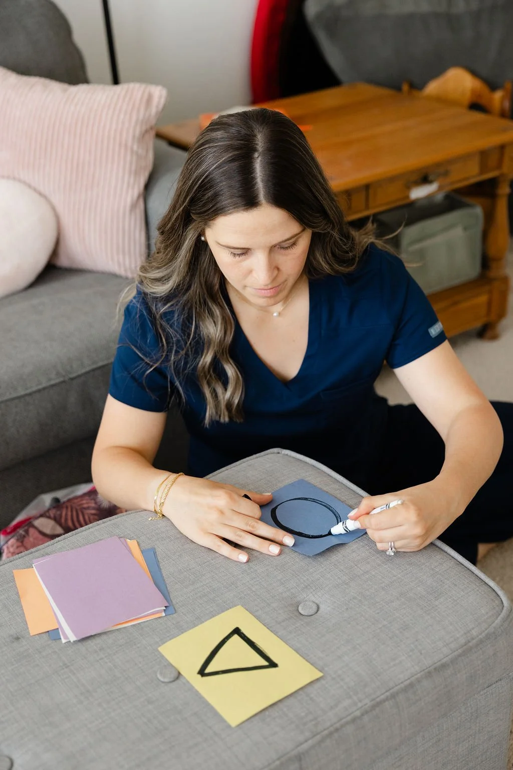 Woman drawing a black shape on a blue paper with a white marker, sitting at a table with colored paper sheets around her in a cozy living room.