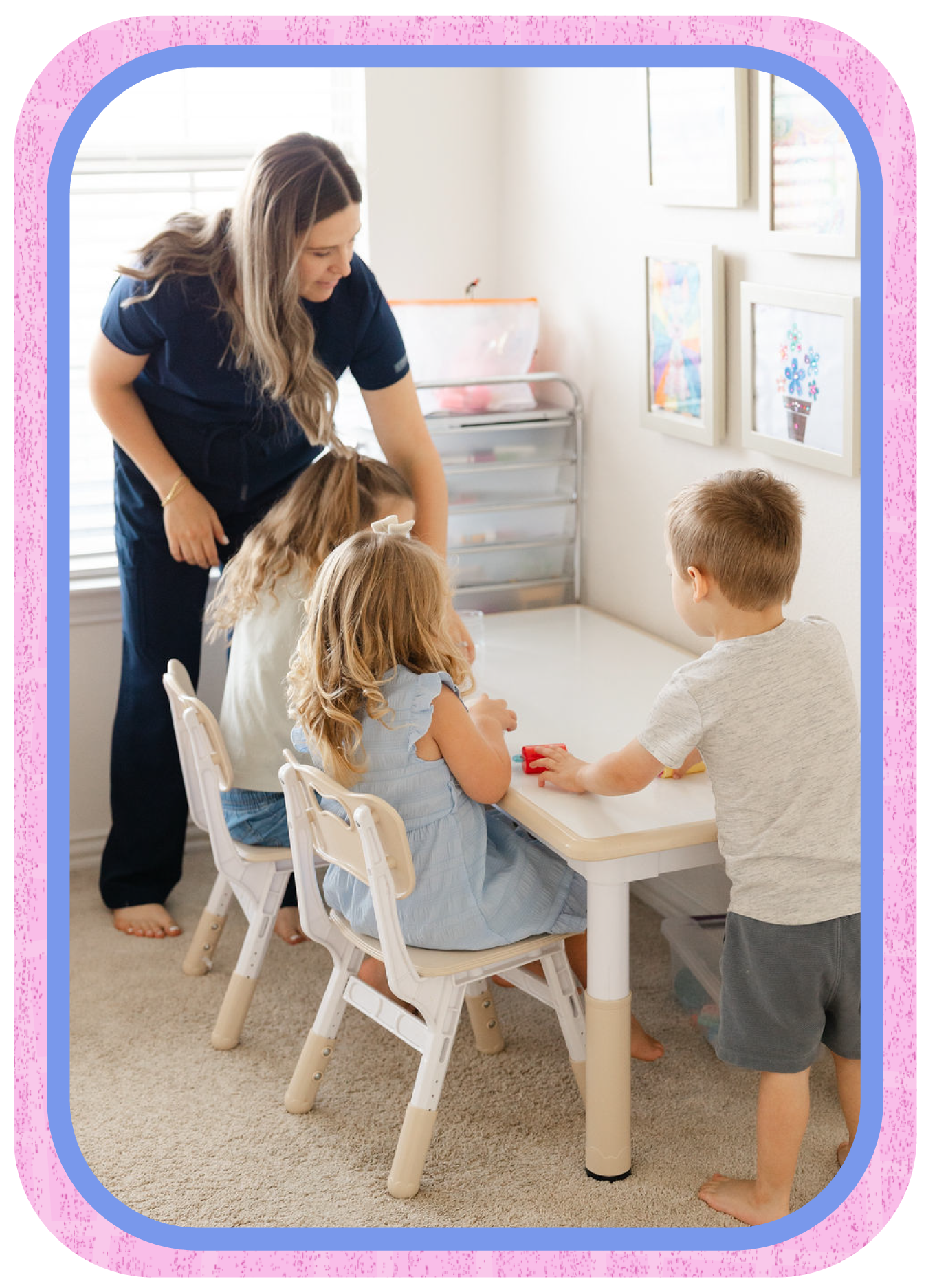 A teacher helping three young children with an activity at a table in a classroom decorated with framed artwork on the wall.
