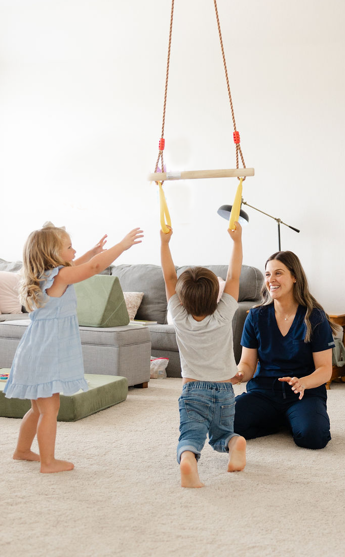 A woman and two children playing indoors with a hanging toy bar. One child is reaching up, and the other is holding a yellow swing attached to the bar.