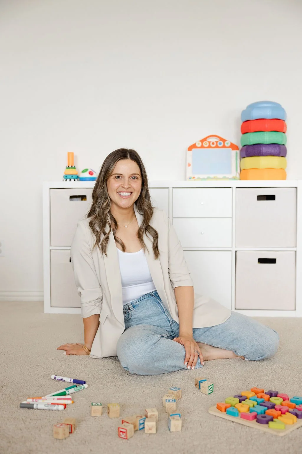 Woman sitting on carpeted floor smiling with children's toys and blocks scattered around, white storage unit with colorful foam blocks and toy on top in background.
