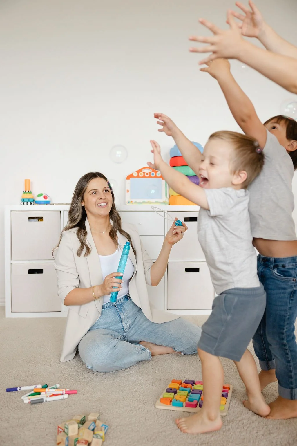 A woman playing with children, blowing bubbles indoors, with some toys on the floor and a colorful toy storage unit in the background.