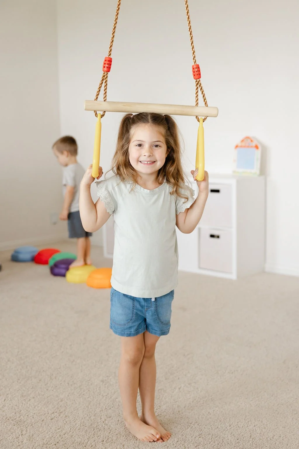 A young girl smiling and holding onto a swing indoors, with a boy in the background stepping over colorful stepping stones.