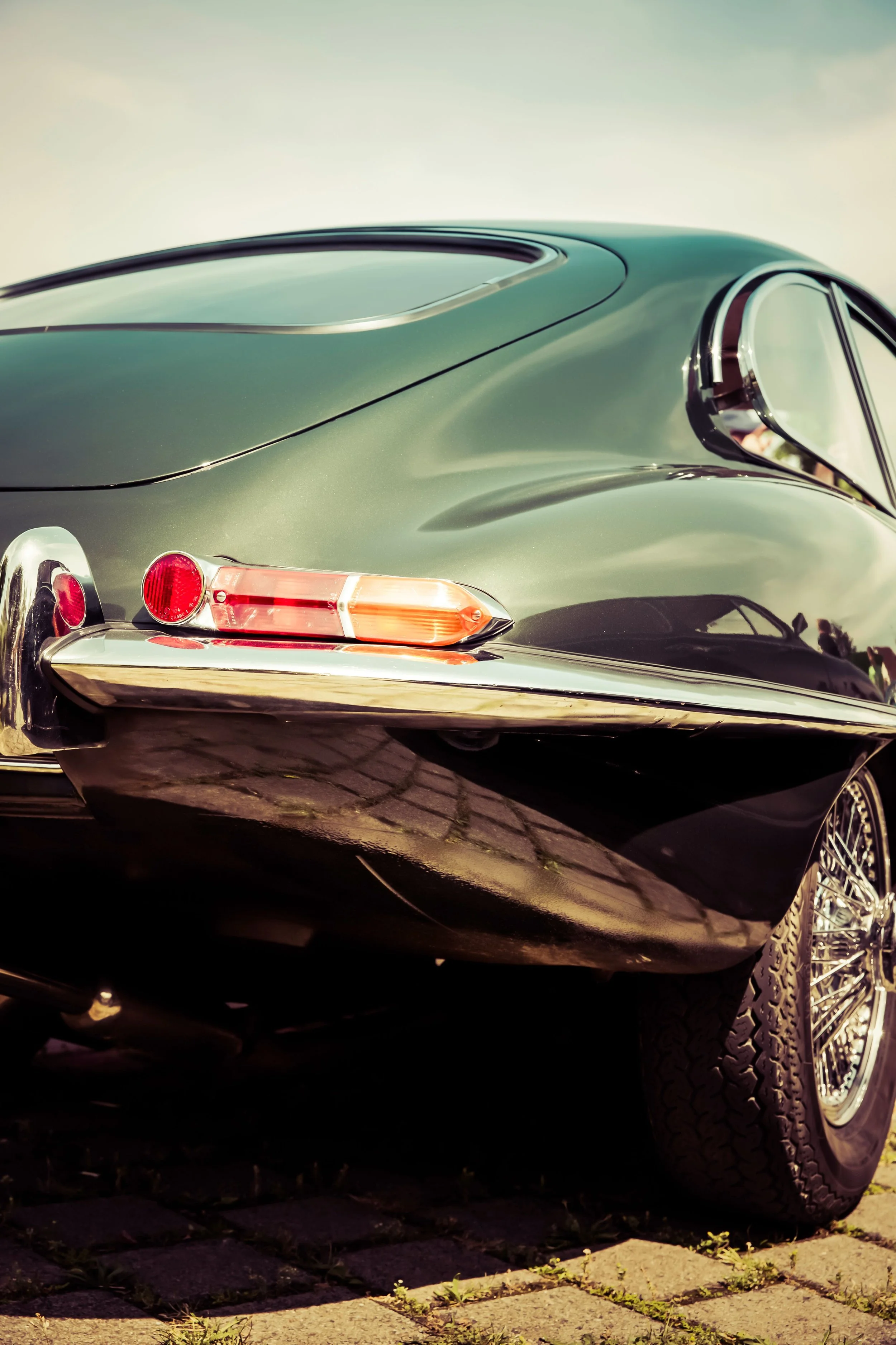 Close-up of the rear side of a vintage dark green car parked on a brick driveway, with a cloudy sky in the background.