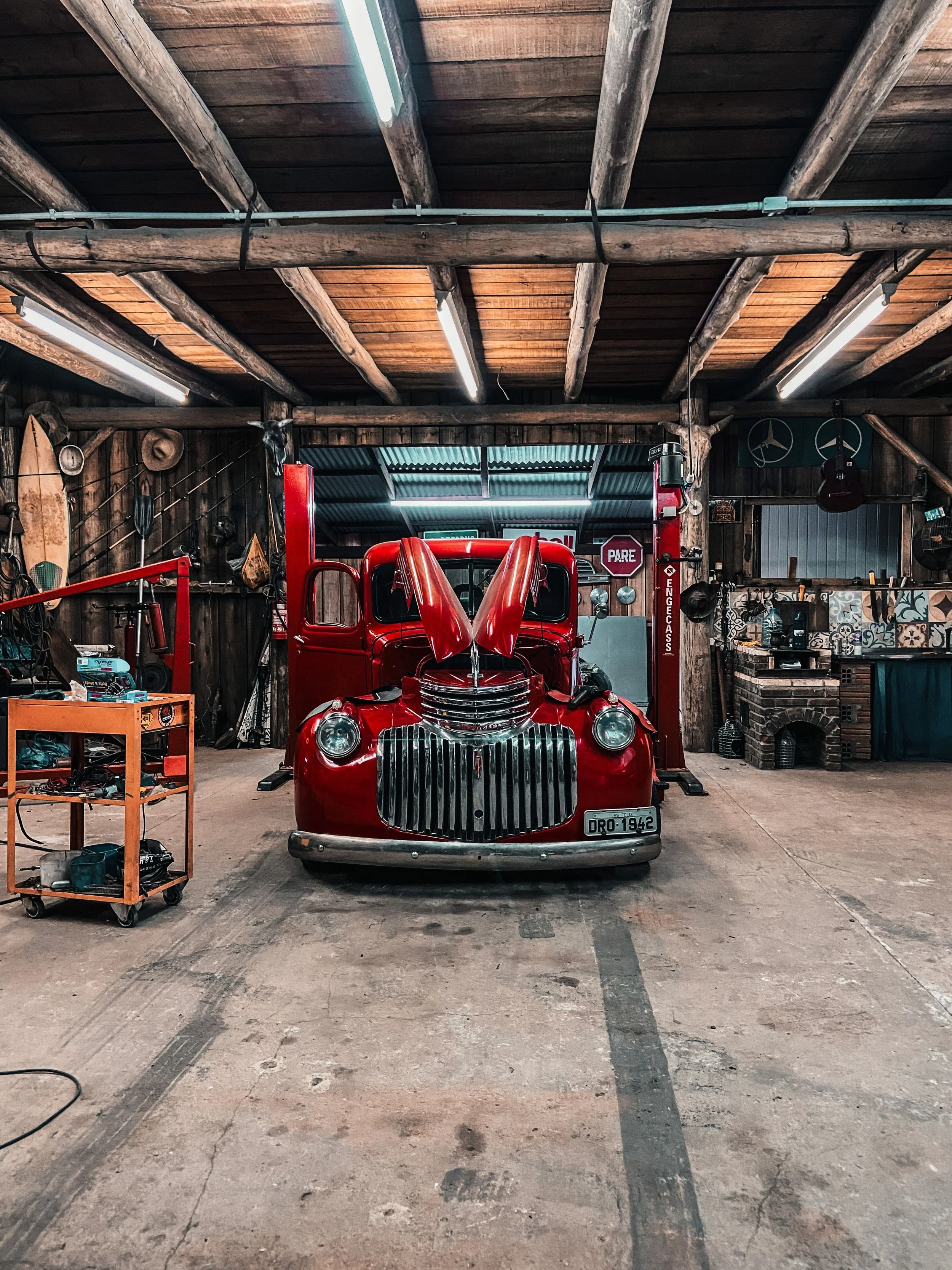 Inside a rustic garage or workshop with wood-paneled walls and ceiling, featuring a vintage red truck with its hood open, surrounded by various tools, equipment, and decorations.
