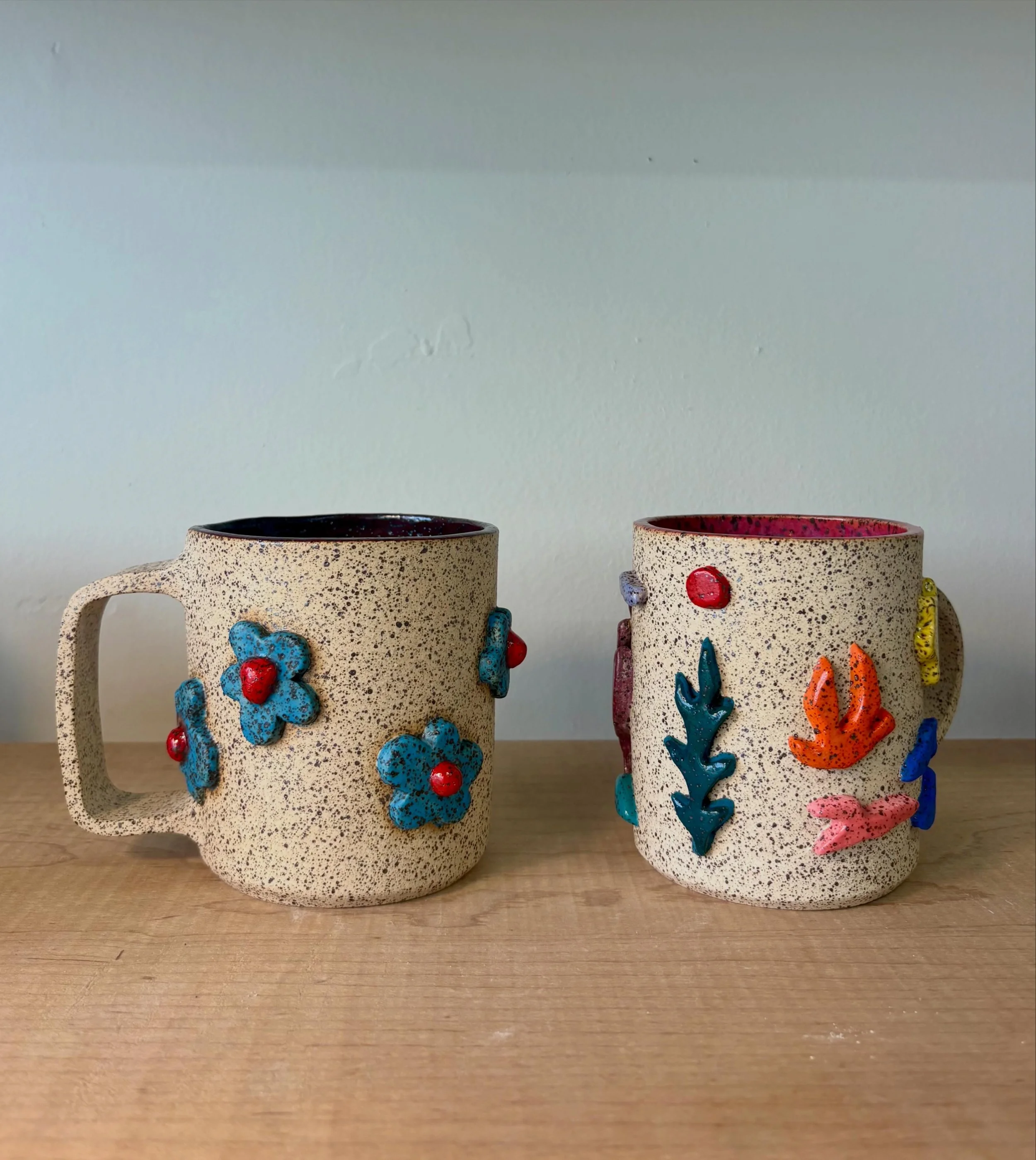 Two ceramic mugs with colorful, textured 3D floral and leaf embellishments, sitting on a wooden surface against a plain wall.