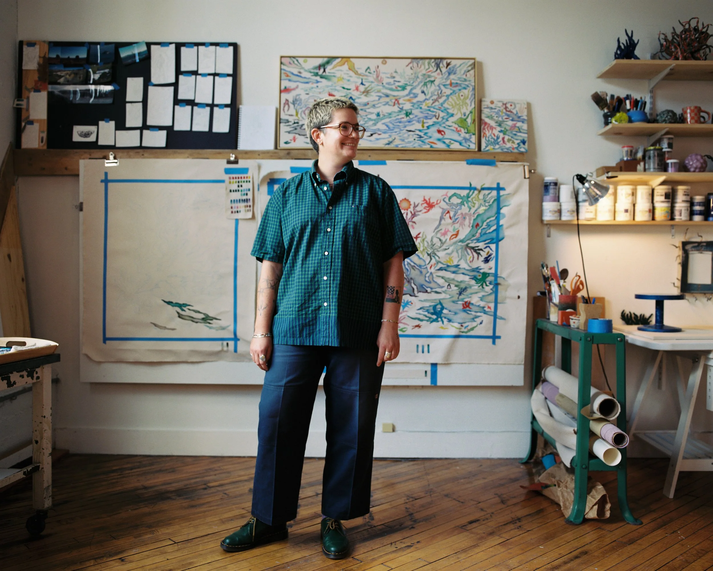 A person with glasses and a checkered shirt standing in an art studio with paintings and supplies in the background.