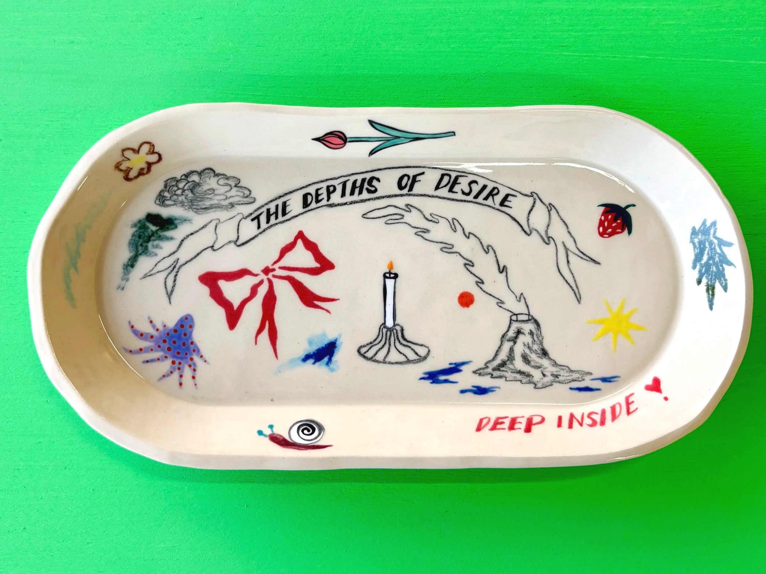 A decorative ceramic tray with various illustrations including a flower, strawberry, candle, mountain, and spiral shell, along with the text 'The depths of desire' and 'Deep inside' on a bright green background.