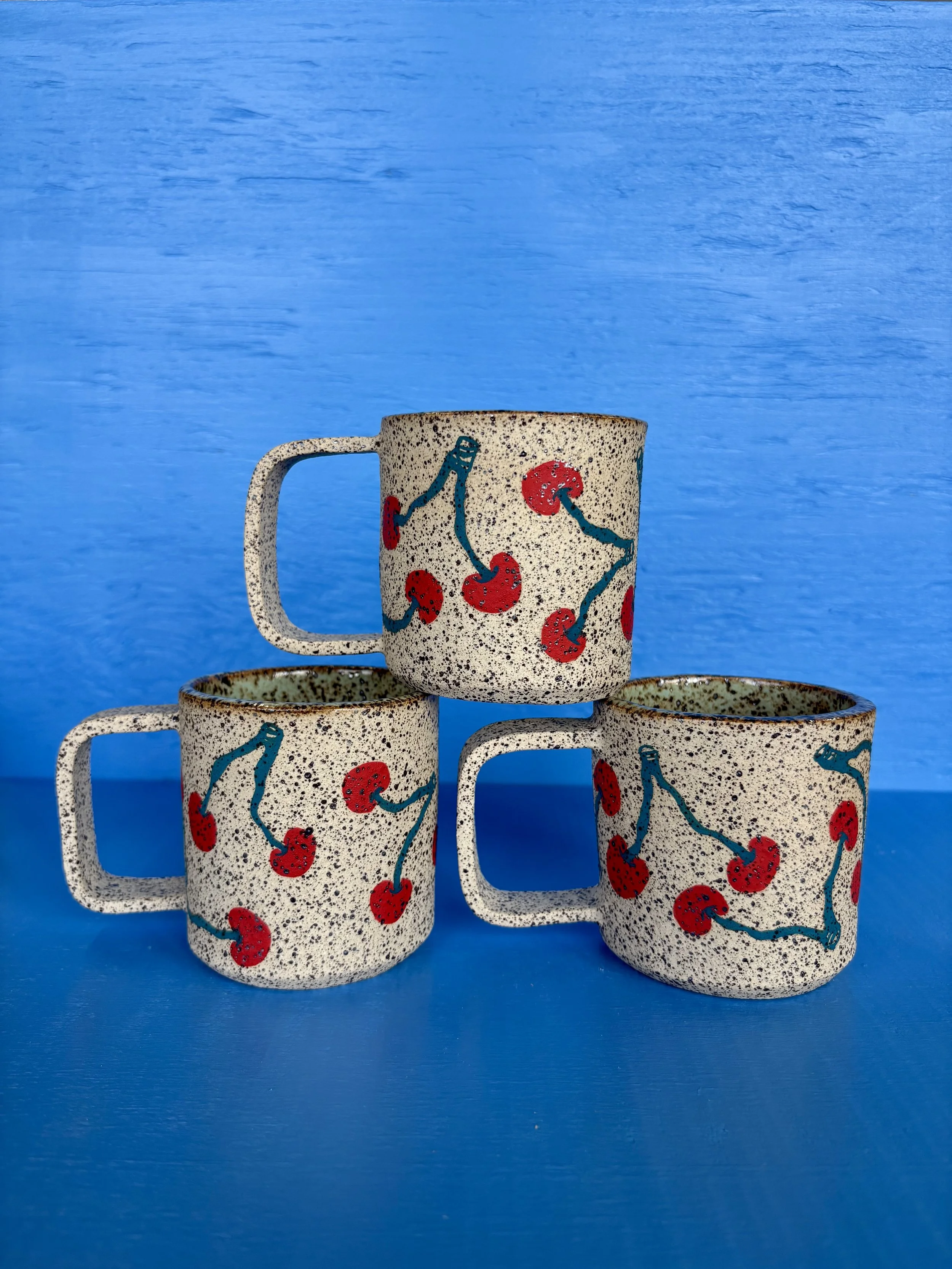 Three speckled ceramic mugs with red cherry and blue vine design, stacked against a bright blue wooden background.