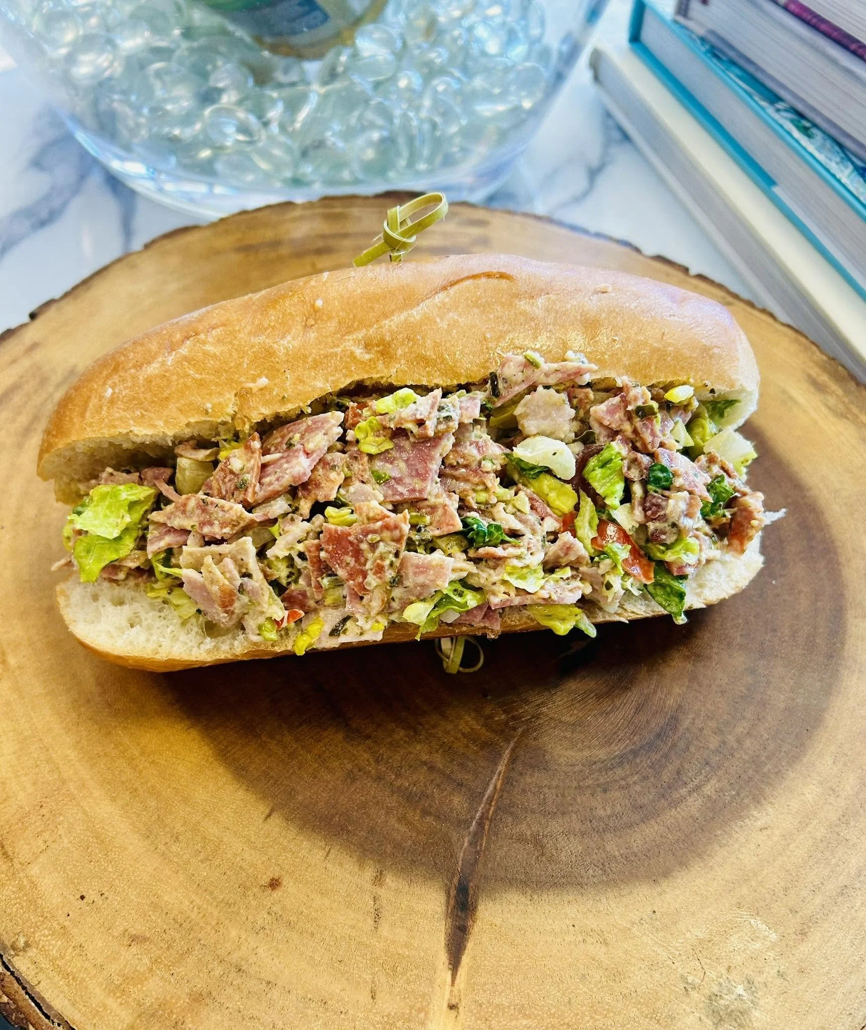 A sandwich with a loaf of bread filled with chopped meat, lettuce, and other vegetables, placed on a wooden surface, with a glass bowl of water with ice and a stack of books in the background.