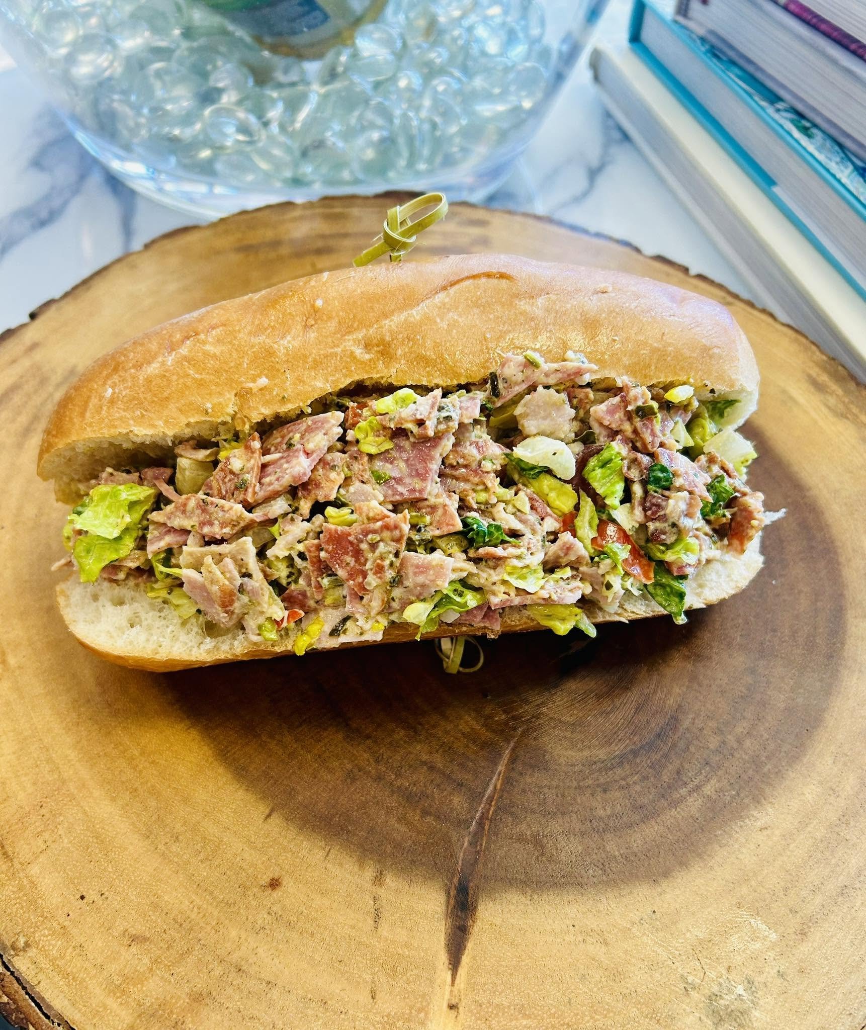 Sandwich with chopped meat and vegetables on a bun, on a wooden cutting board, with a glass container of water and books in the background.