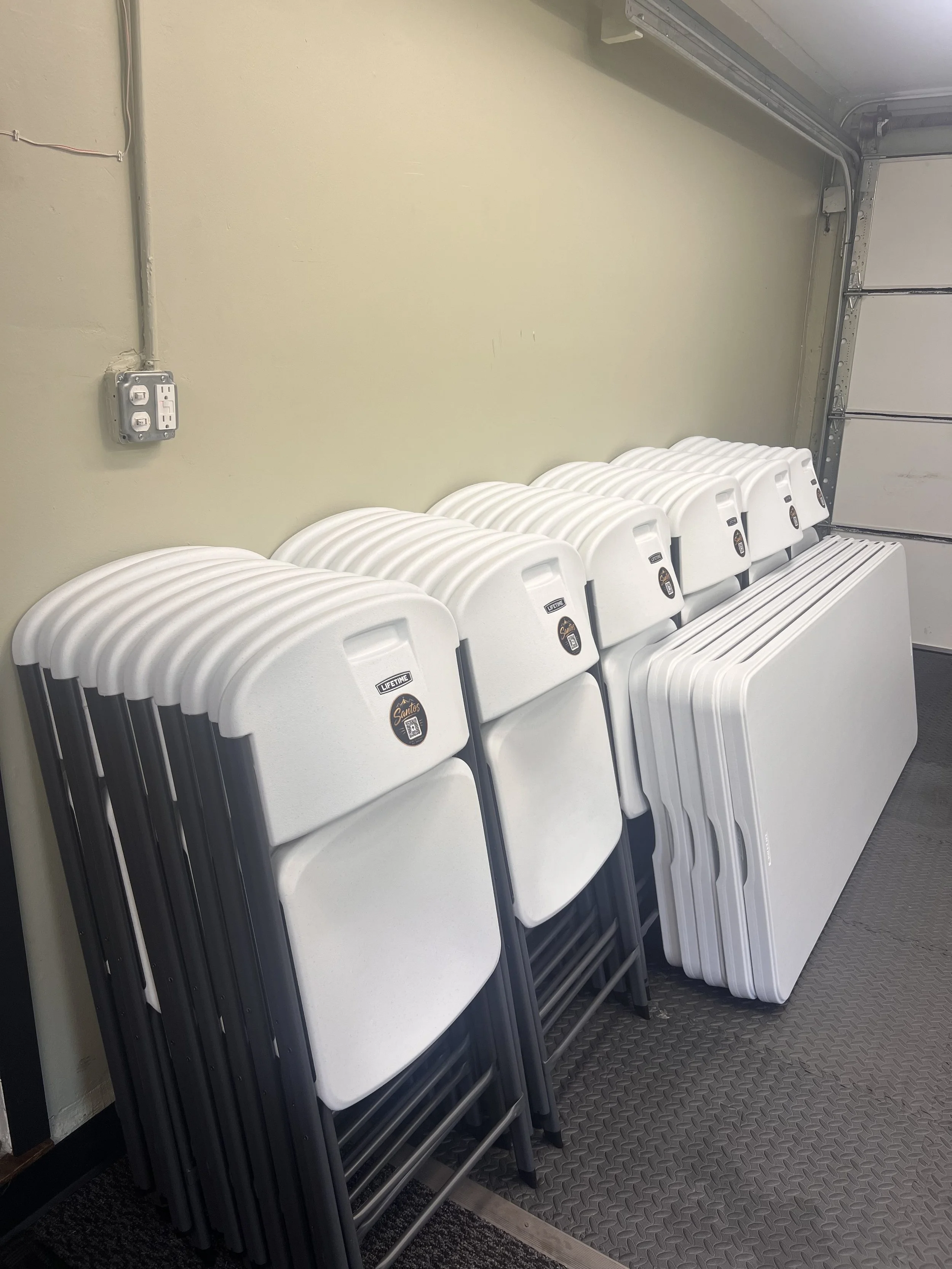 Stacked white folding chairs with black legs placed against a pale yellow wall in a garage or storage room. There is a closed garage door on the right side and an electrical outlet with a socket and switch on the wall.