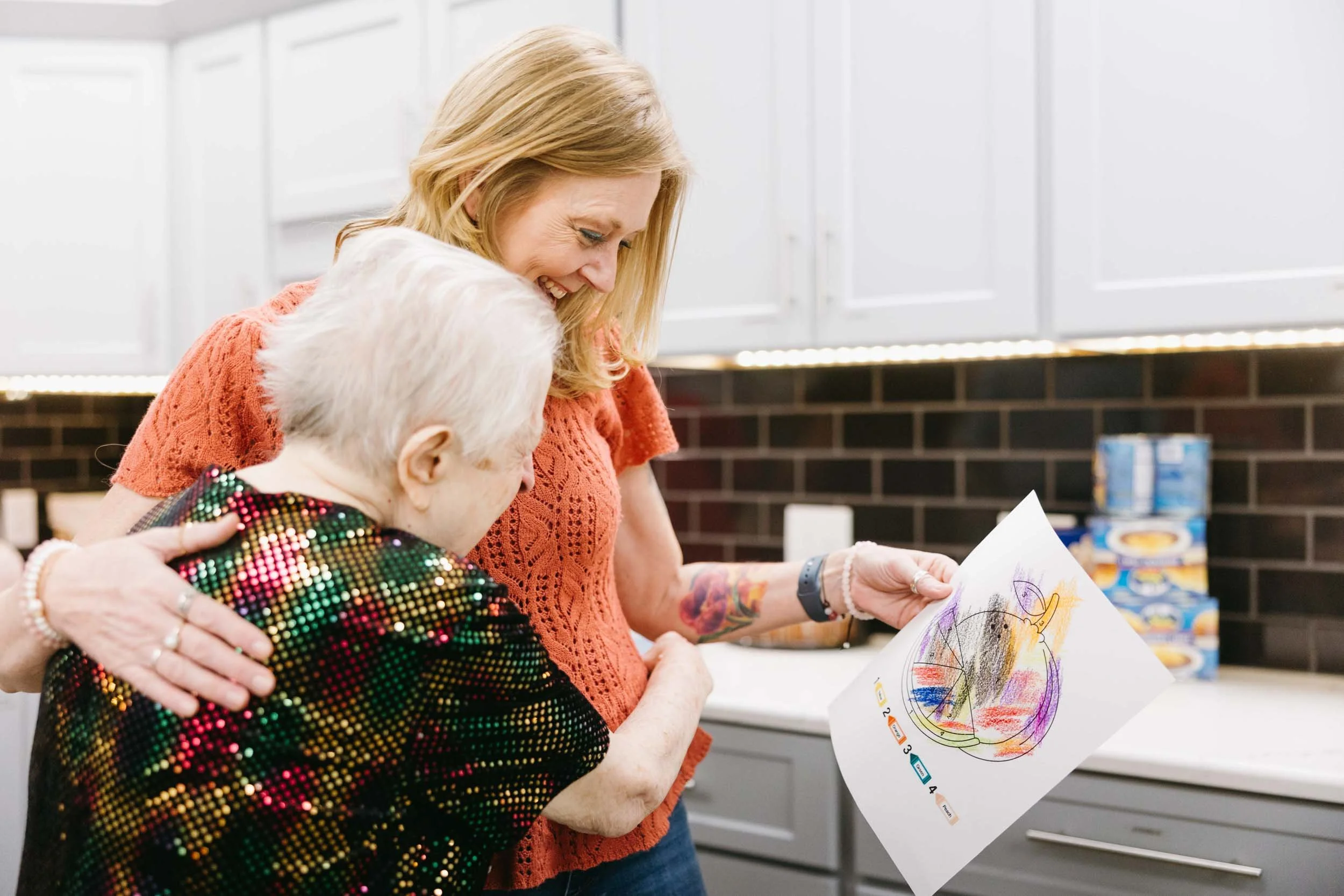 Women sharing an artwork that has colorful circular scribbles, in a kitchen setting.