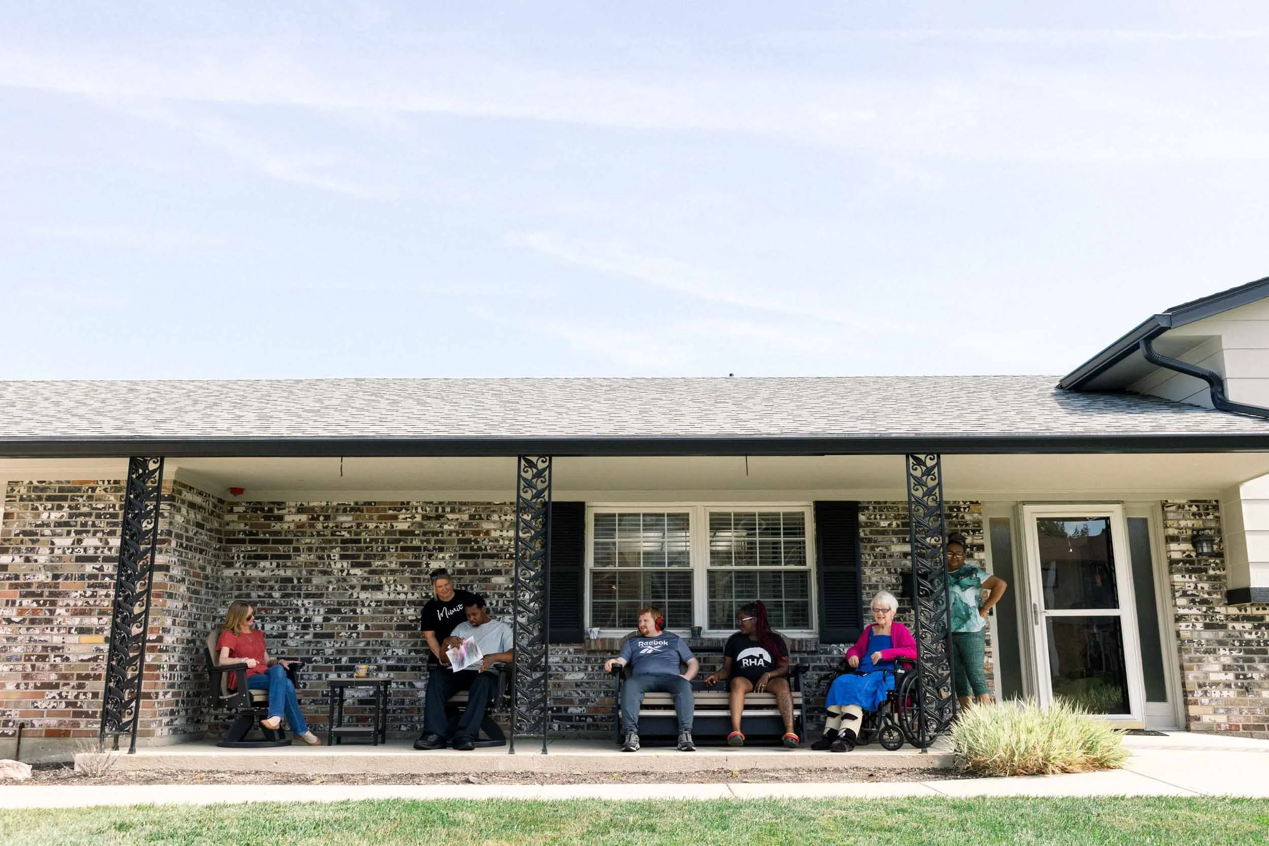 People sitting and standing on a covered porch, socializing, with a brick house in the background.