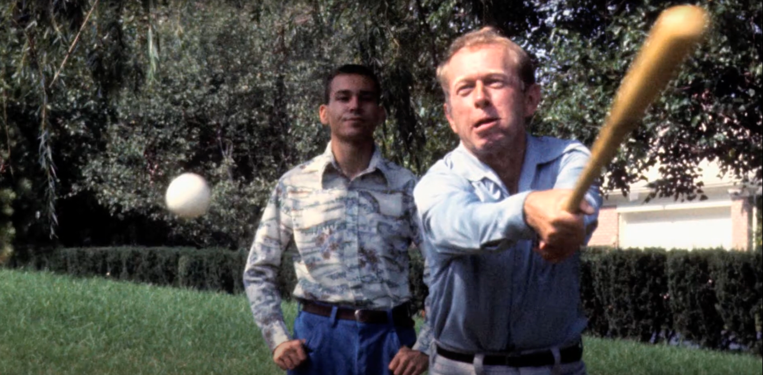 Two men playing golf outdoors, one swinging a golf club while the other observes, with trees and a house in the background.