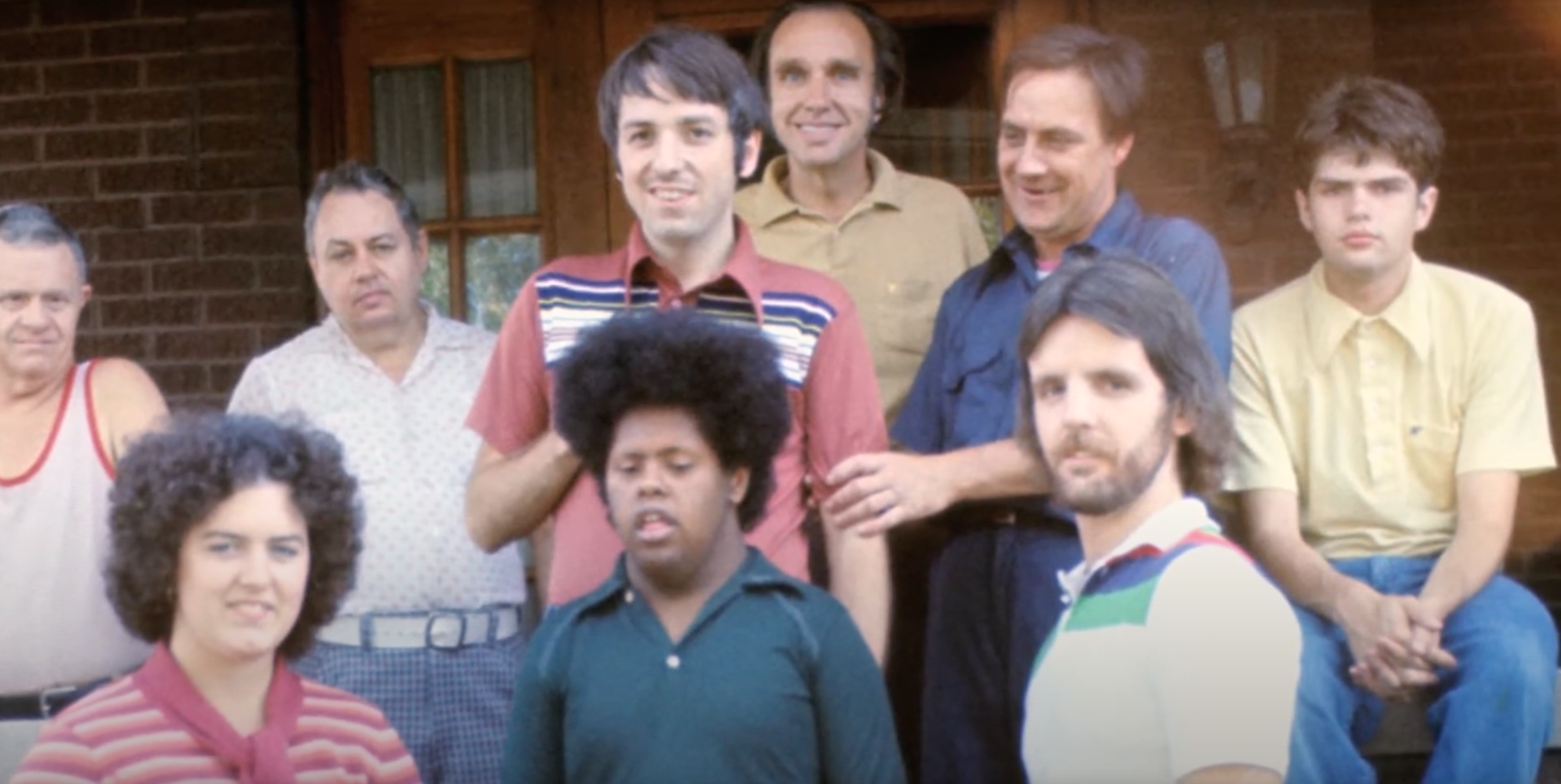 A group of people standing and sitting outside in front of a brick house with wooden window frames, posing for a photo, from the TV show 'The Wonder Years'.