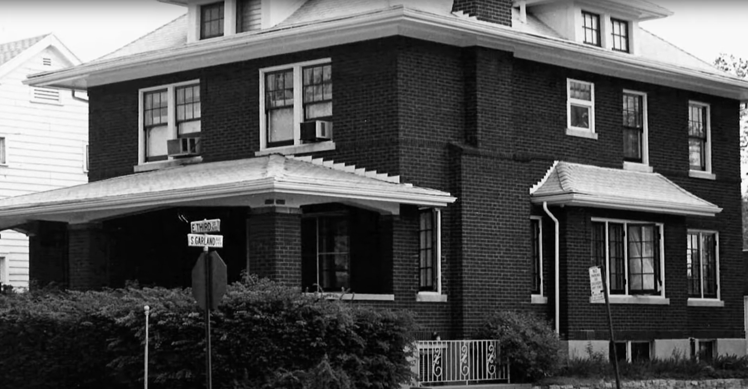 A black and white photo of a three-story brick building at the corner of E. Third Street and S. Garland Avenue, with house signs and a landscaped front yard.