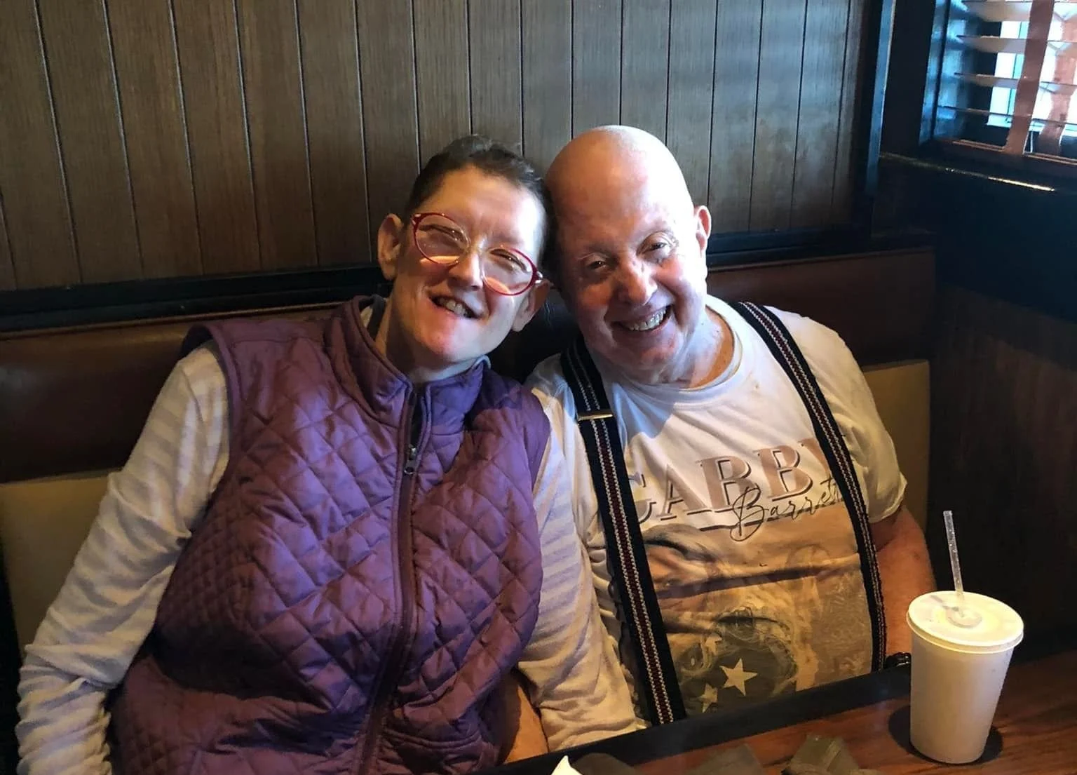 A woman with short dark hair, glasses, and a purple quilted vest, and a bald man with suspenders and a graphic T-shirt, smiling and sitting close together in a restaurant booth.