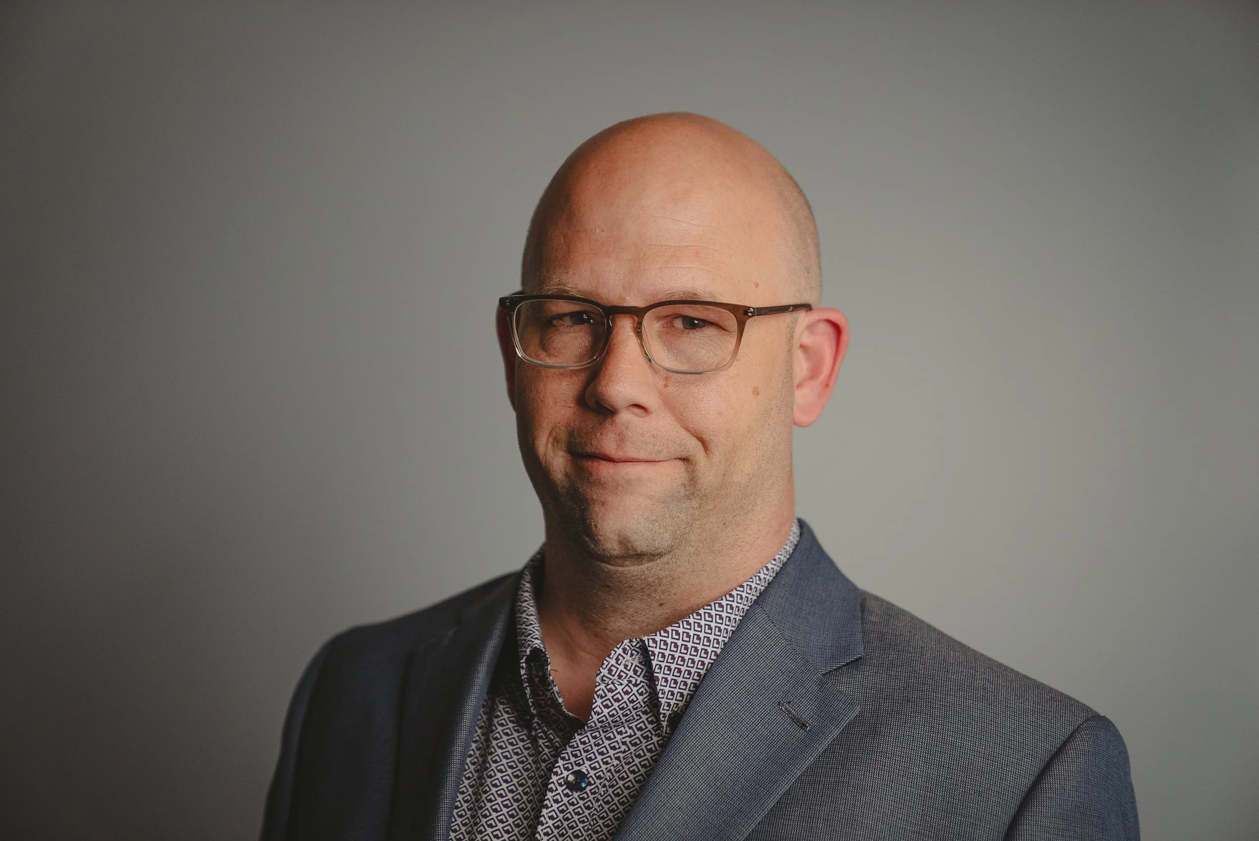 A middle-aged man with glasses, a bald head, wearing a patterned dress shirt and a dark blazer, smiling slightly, against a plain gray background.