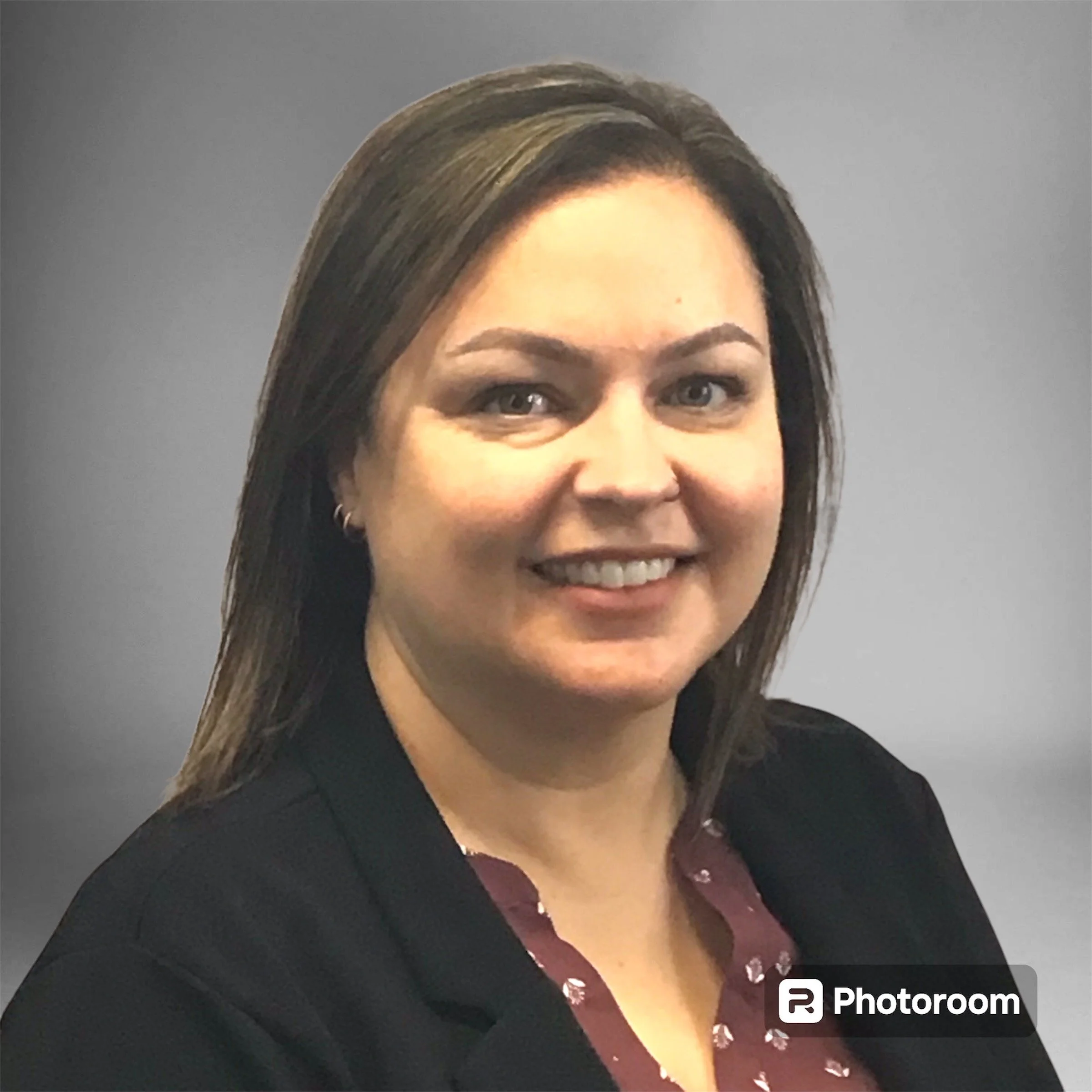 Close-up photo of a smiling woman with light skin, long dark hair with highlights, wearing a black blazer and a maroon shirt with small light-colored patterns. The background is plain gray.
