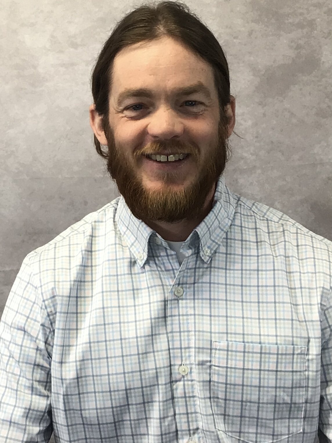 A man with brown hair, a beard, and a mustache is smiling, wearing a light-colored checkered shirt, standing against a plain gray background.