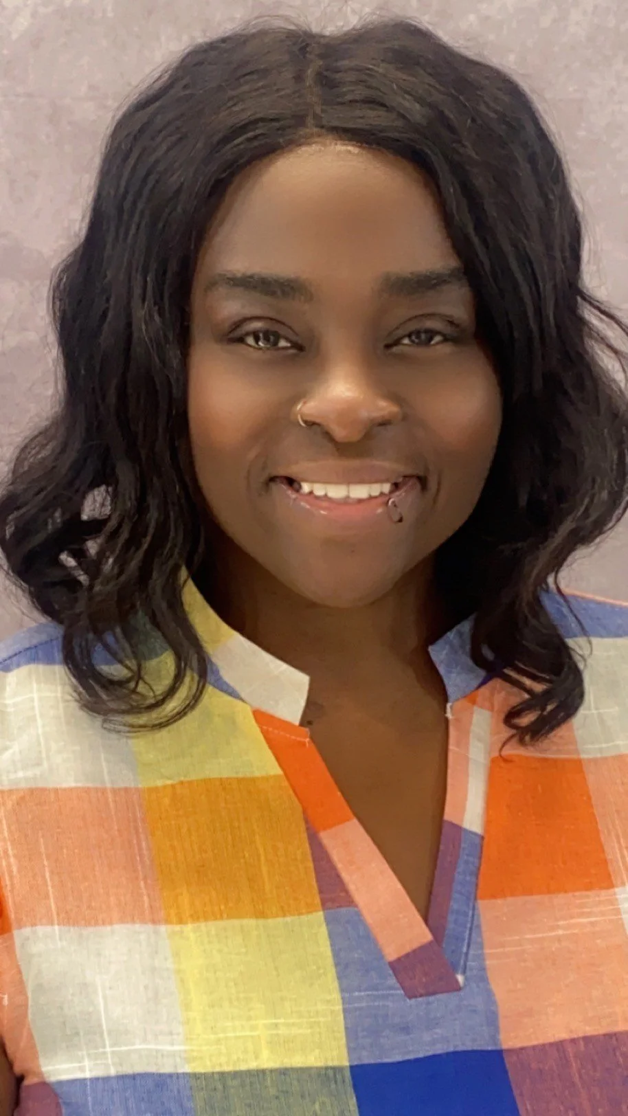 A young woman with dark wavy hair, wearing a multicolored shirt and a nose ring, smiling at the camera.