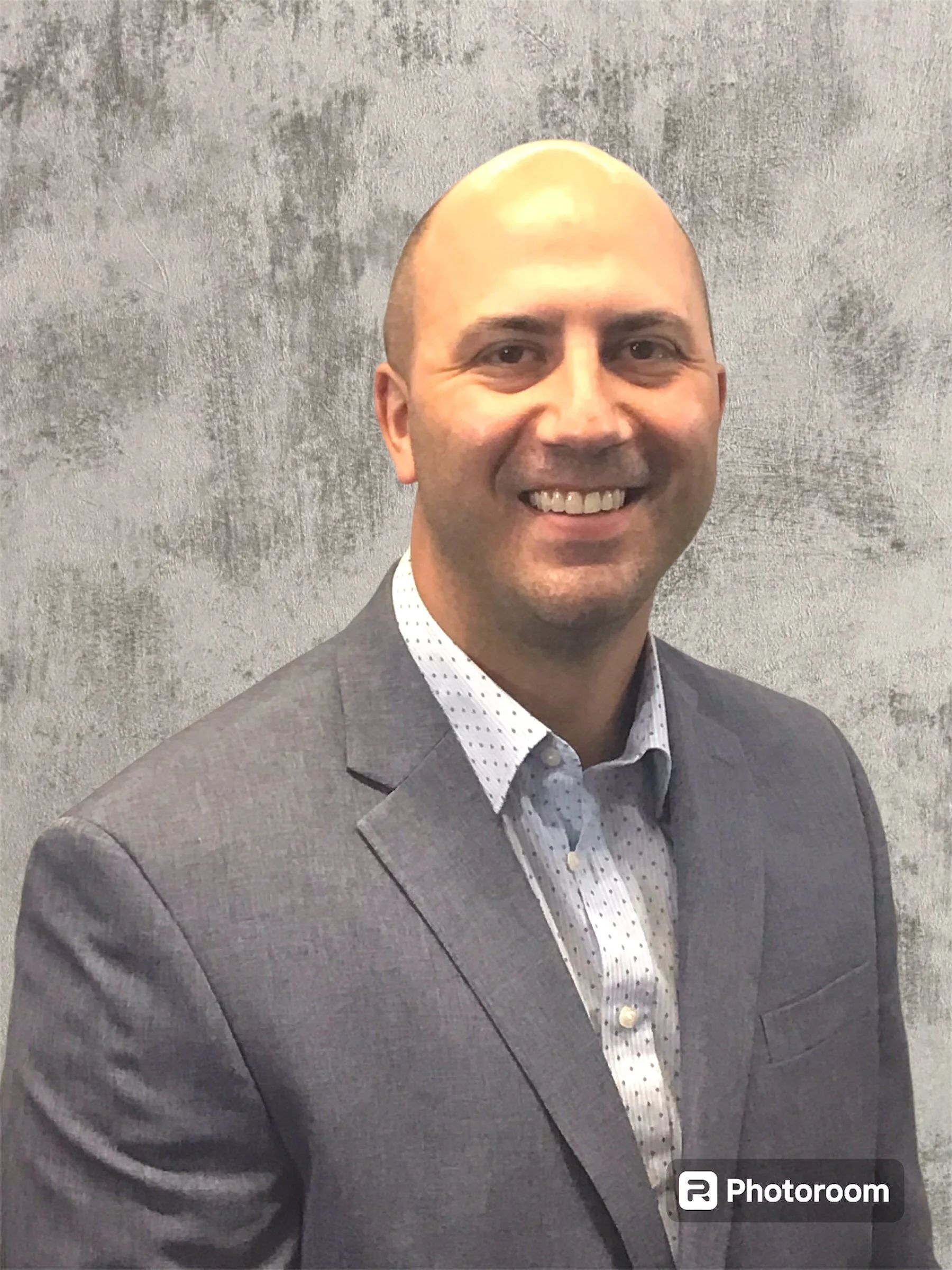 A smiling man in a grey suit and white shirt standing against a grey textured wall.