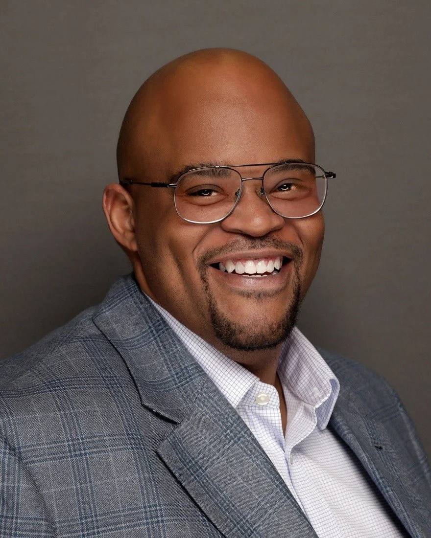 A smiling man with a bald head, glasses, and a goatee, wearing a gray checkered blazer and white shirt, against a plain gray background.