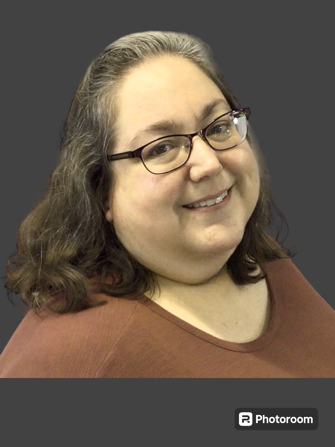 A woman with shoulder-length brown hair, glasses, and a warm smile, wearing a brown top, against a gray background.