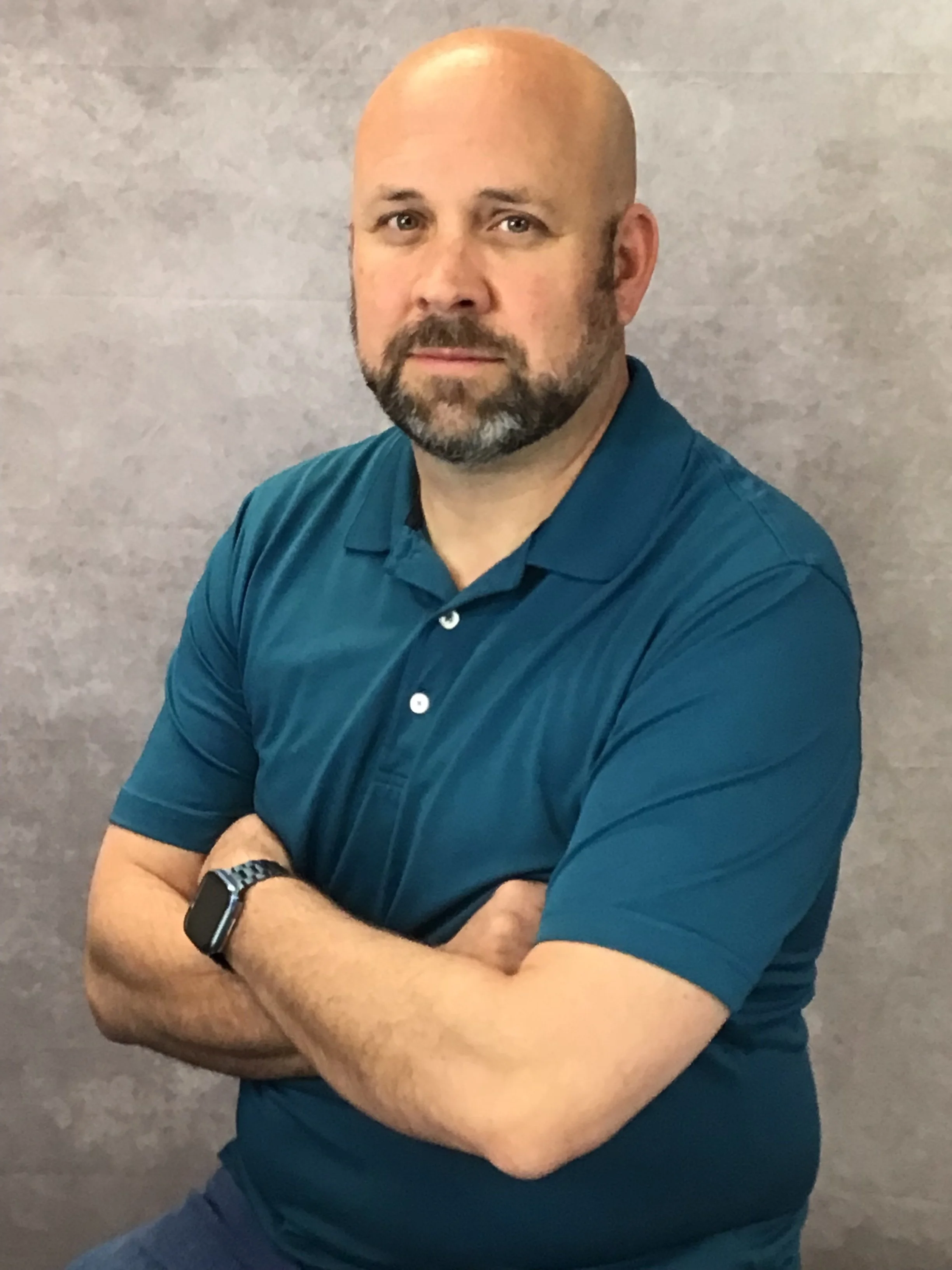 A man with a beard, mustache, and shaved head, wearing a blue polo shirt and a watch, sitting with arms crossed in front of a grey textured wall.