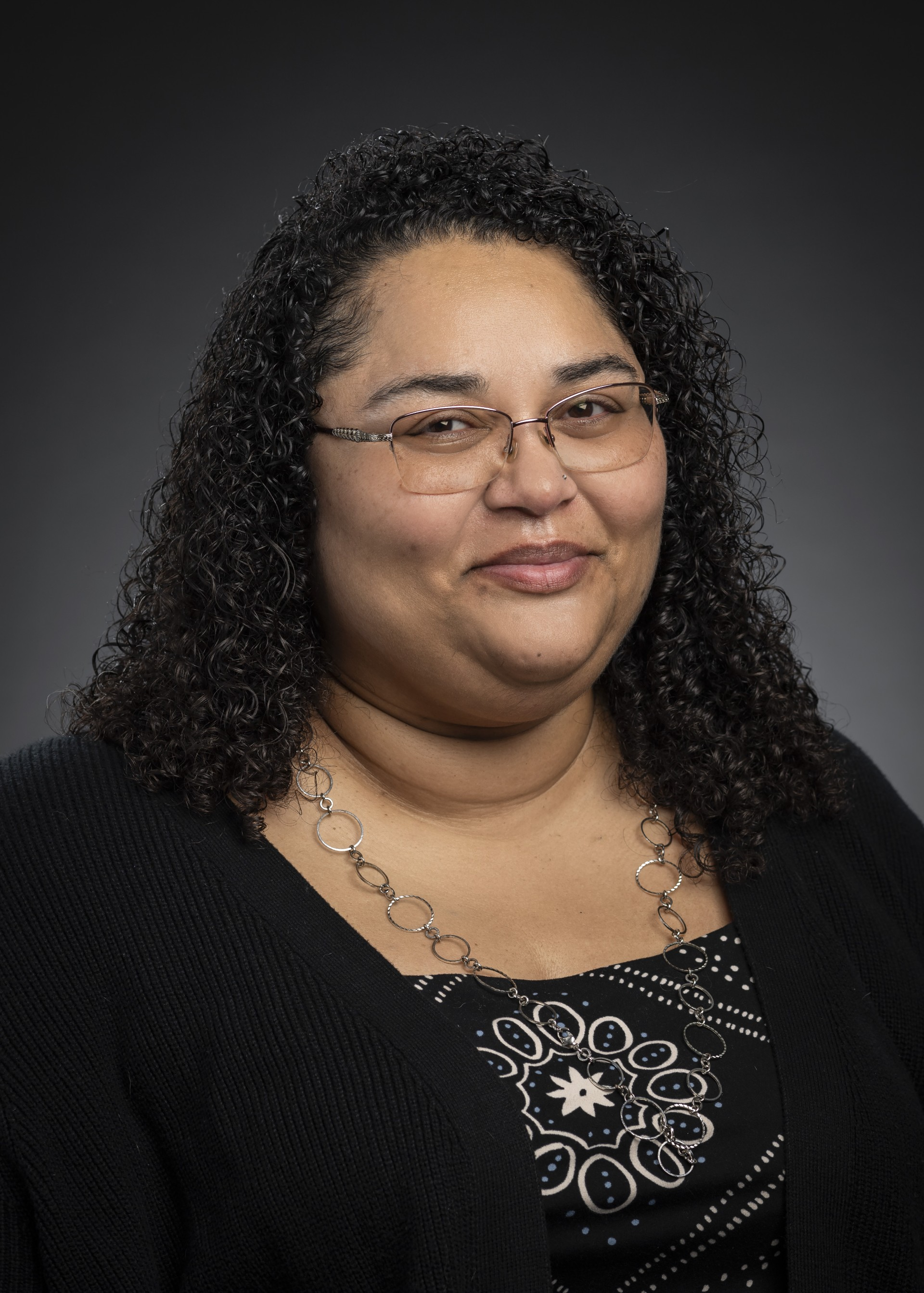 Portrait of a woman with curly black hair, wearing glasses, a black top with a patterned shirt underneath, a silver necklace, and a friendly expression against a dark background.