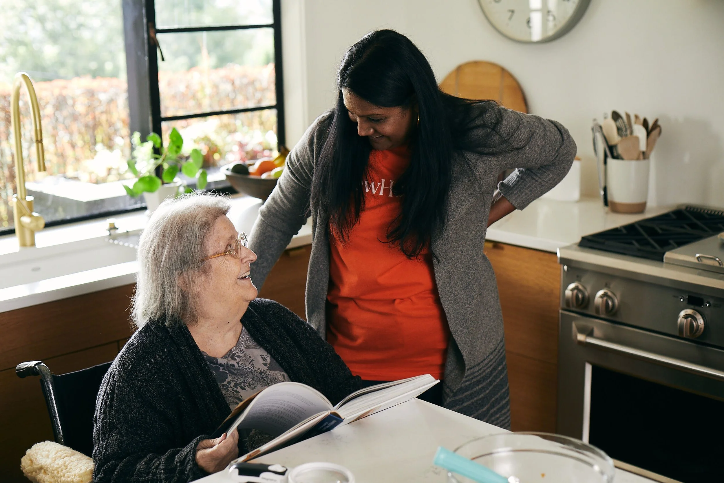 An elderly woman in a wheelchair reading a book and smiling at a younger woman, likely her caretaker, who is leaning over her, smiling and engaging with her in a kitchen.