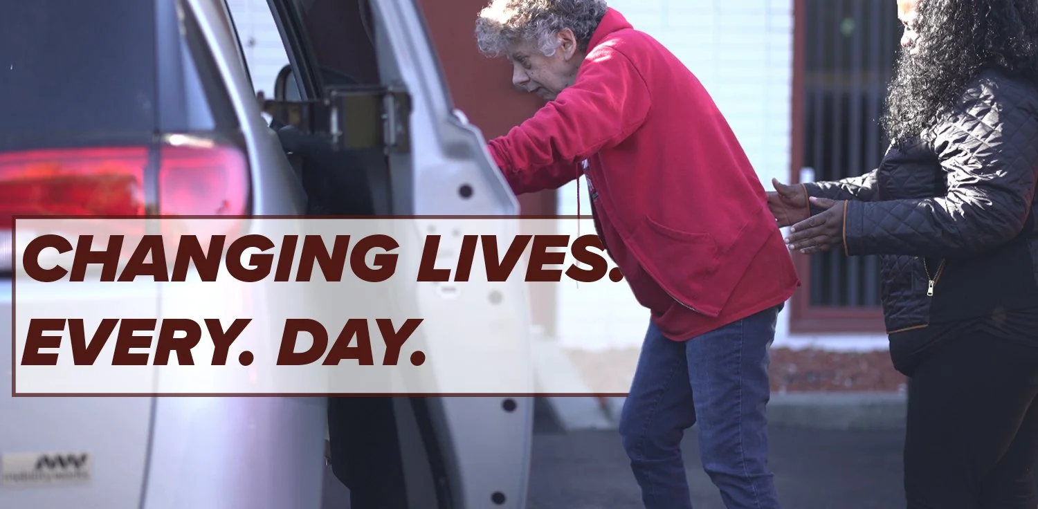 An elderly man with gray curly hair is being assisted by a woman with curly black hair to get into an SUV. The man is wearing a red jacket, and the woman is wearing a black quilted jacket.