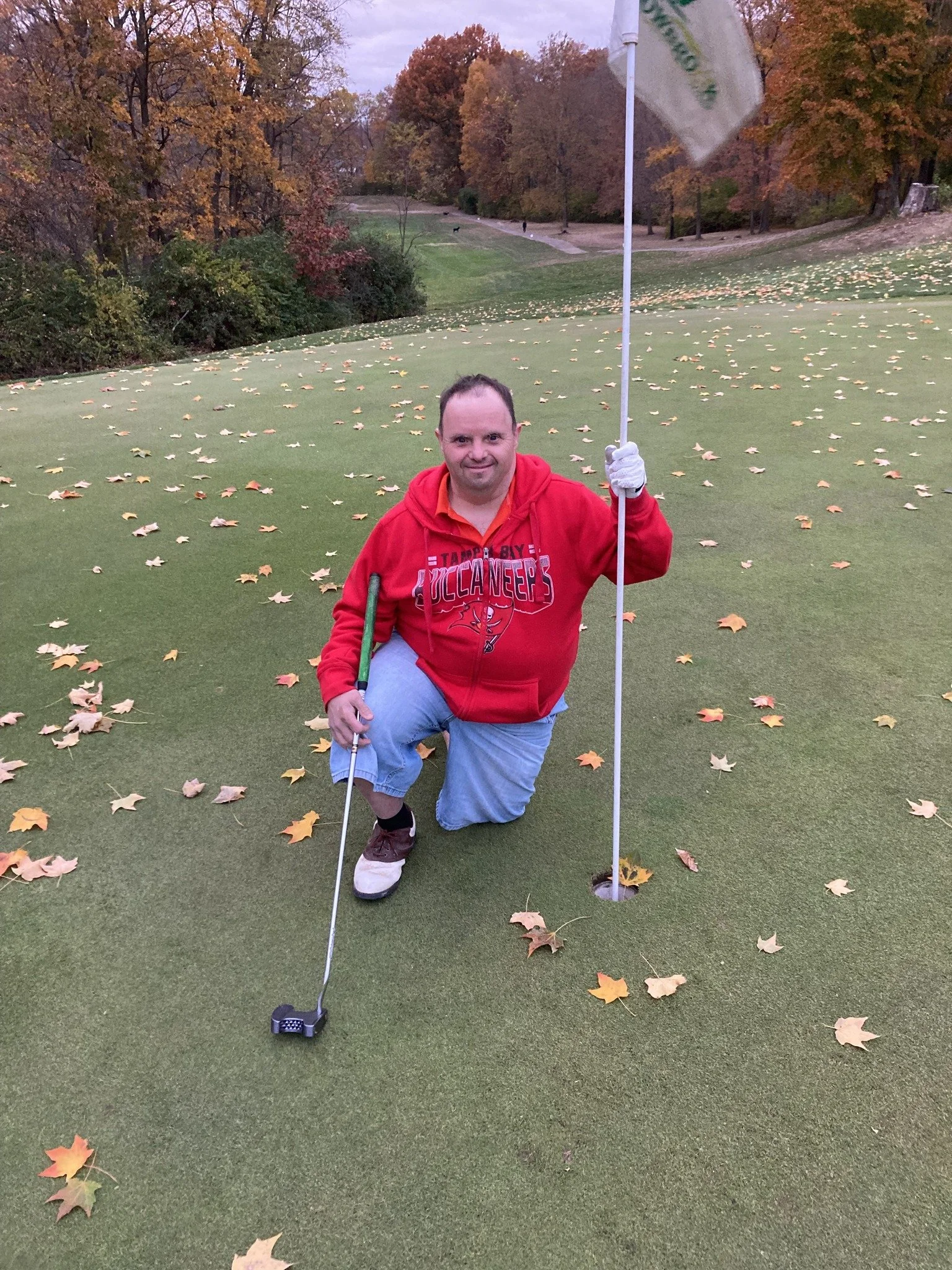 A man in a red hoodie and gray shorts kneeling on a golf course green, holding a golf club and a flagstick, surrounded by fallen autumn leaves with trees in fall colors in the background.