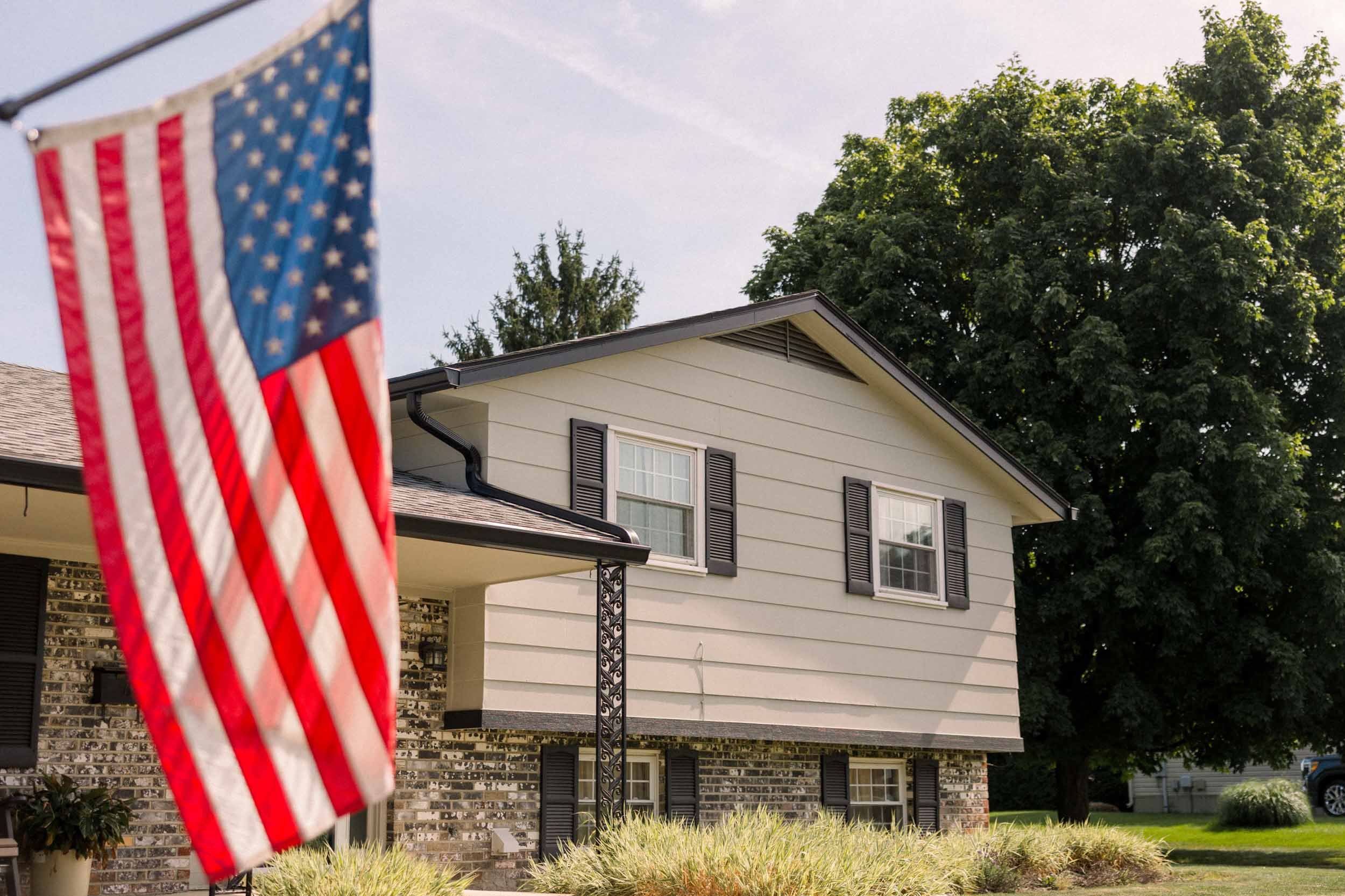American flag hanging in front of a two-story house with beige siding, black shutters, and a brick lower facade, surrounded by green trees and grass.