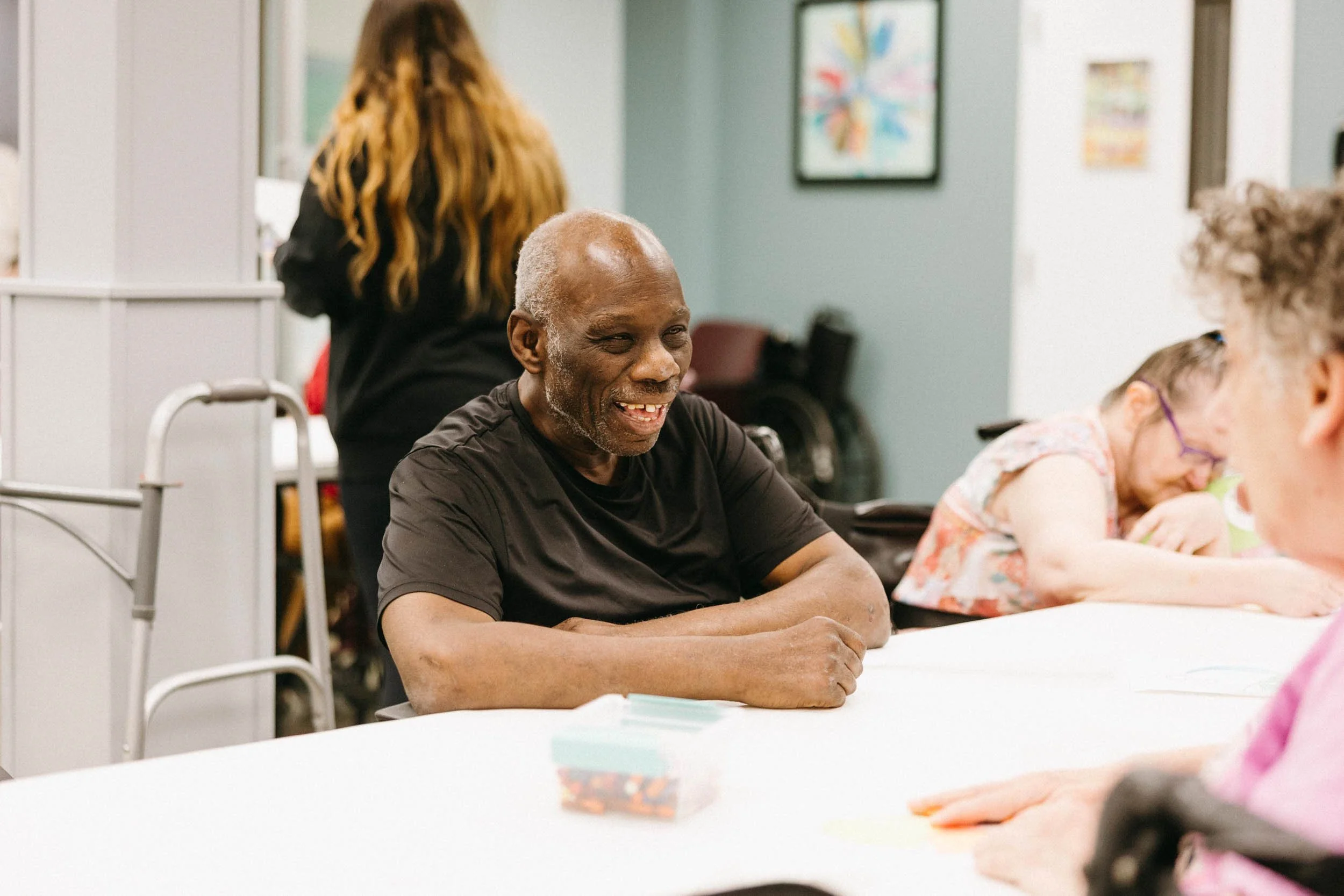 An elderly man smiling and sitting at a table in a group setting, with other children and possibly staff present, engaging in an activity.