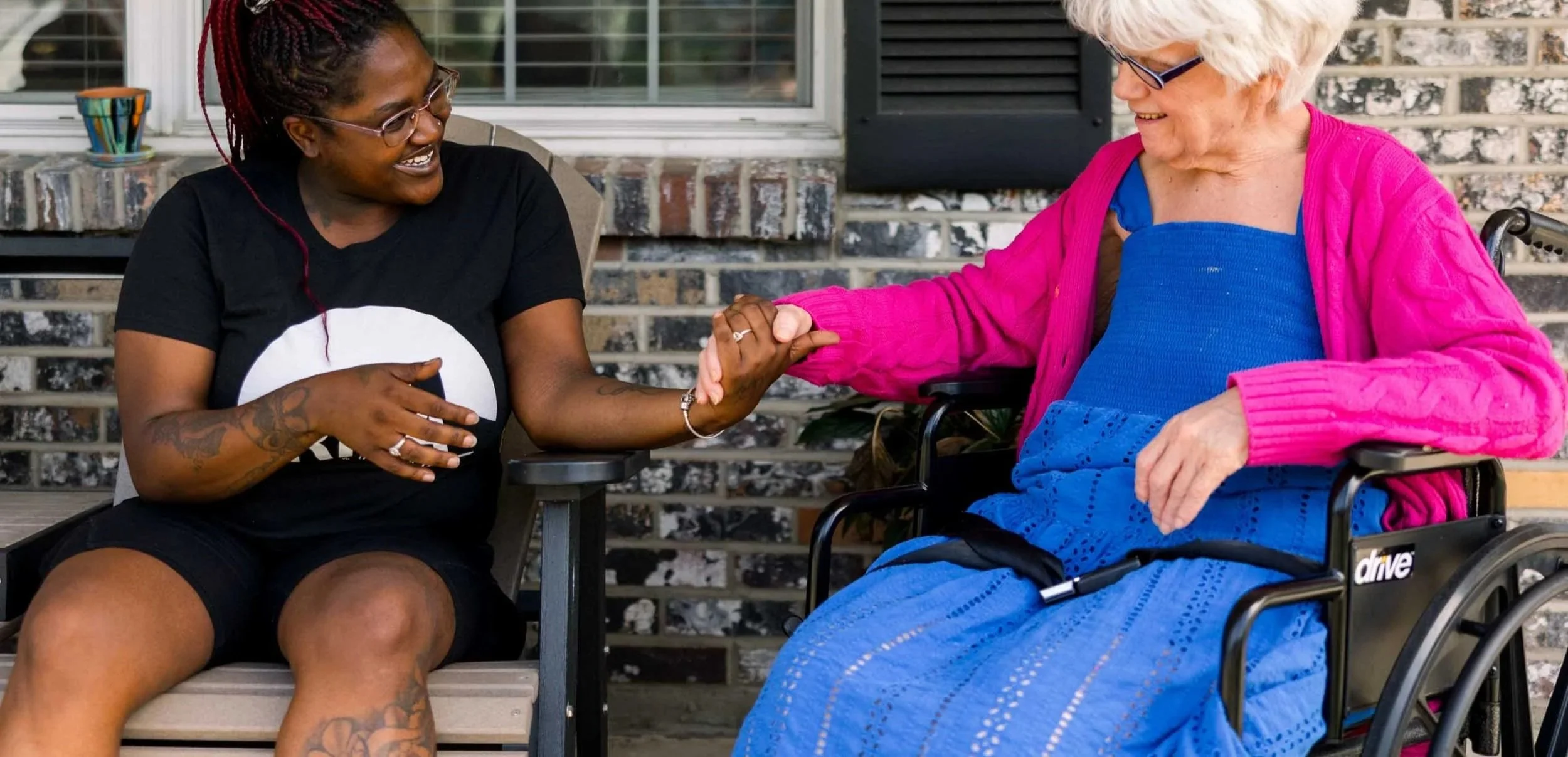 Two women shaking hands, one young with tattoos and glasses, the other elderly in a wheelchair with white hair and glasses, smiling and engaging in an outdoor setting.