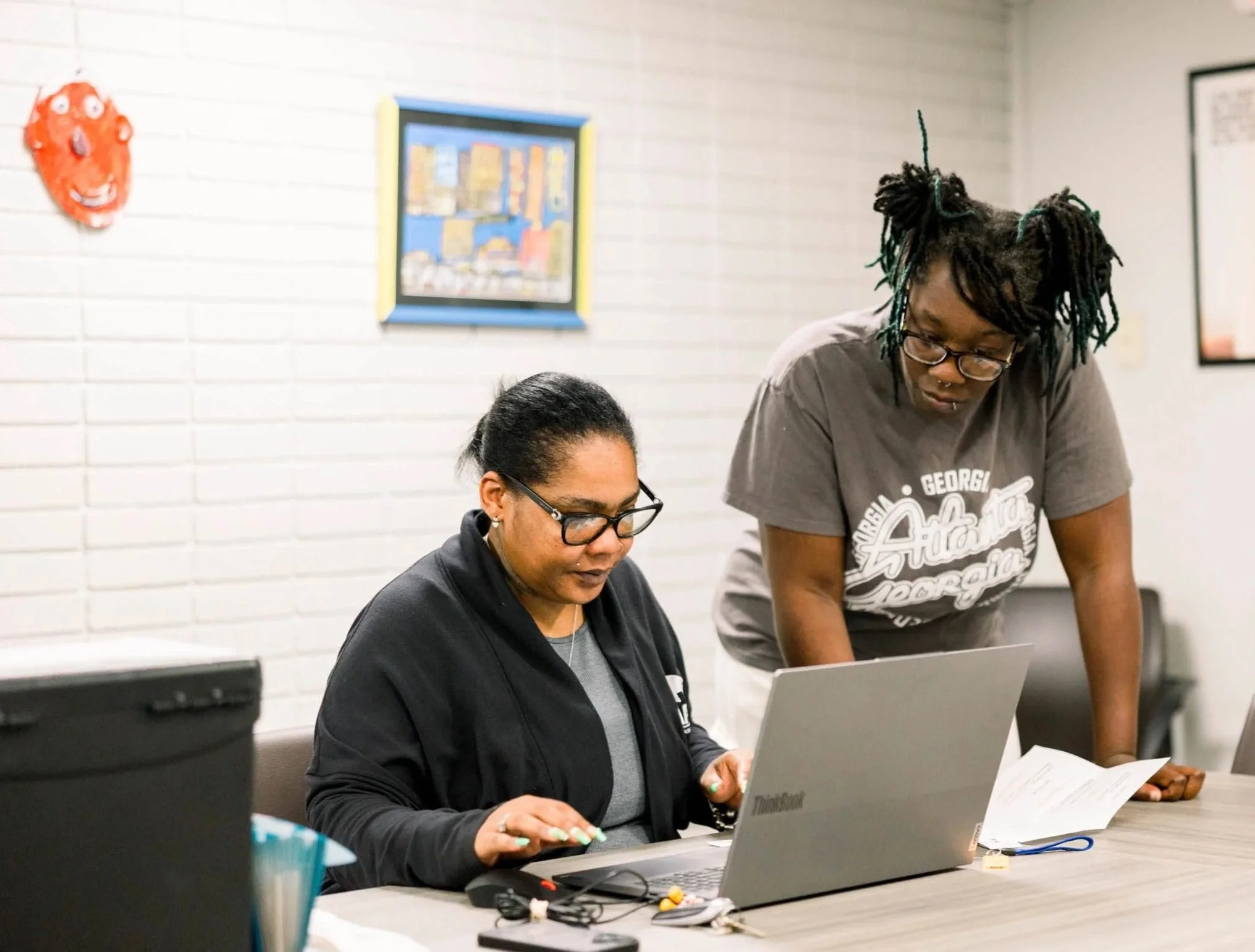 Two women are working together at a table with a laptop and papers, in an indoor setting with white brick walls and framed artwork on the wall.