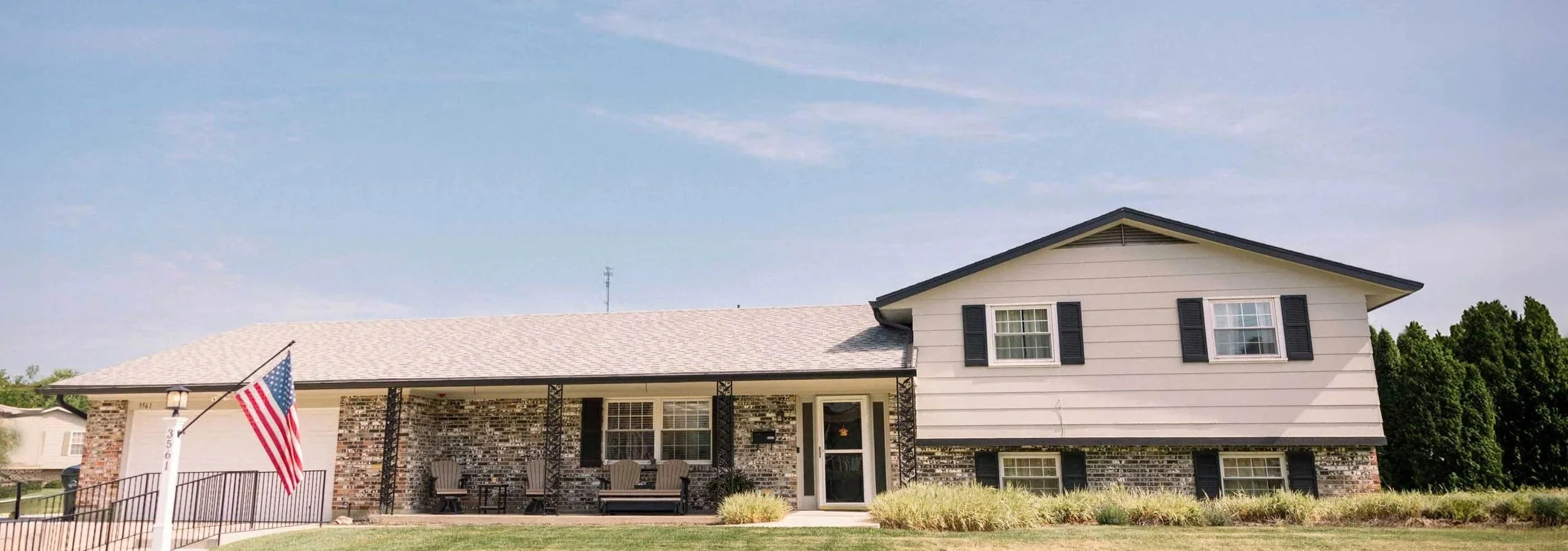A two-story house with beige siding and black shutters, front porch with chairs, and an American flag in the yard.
