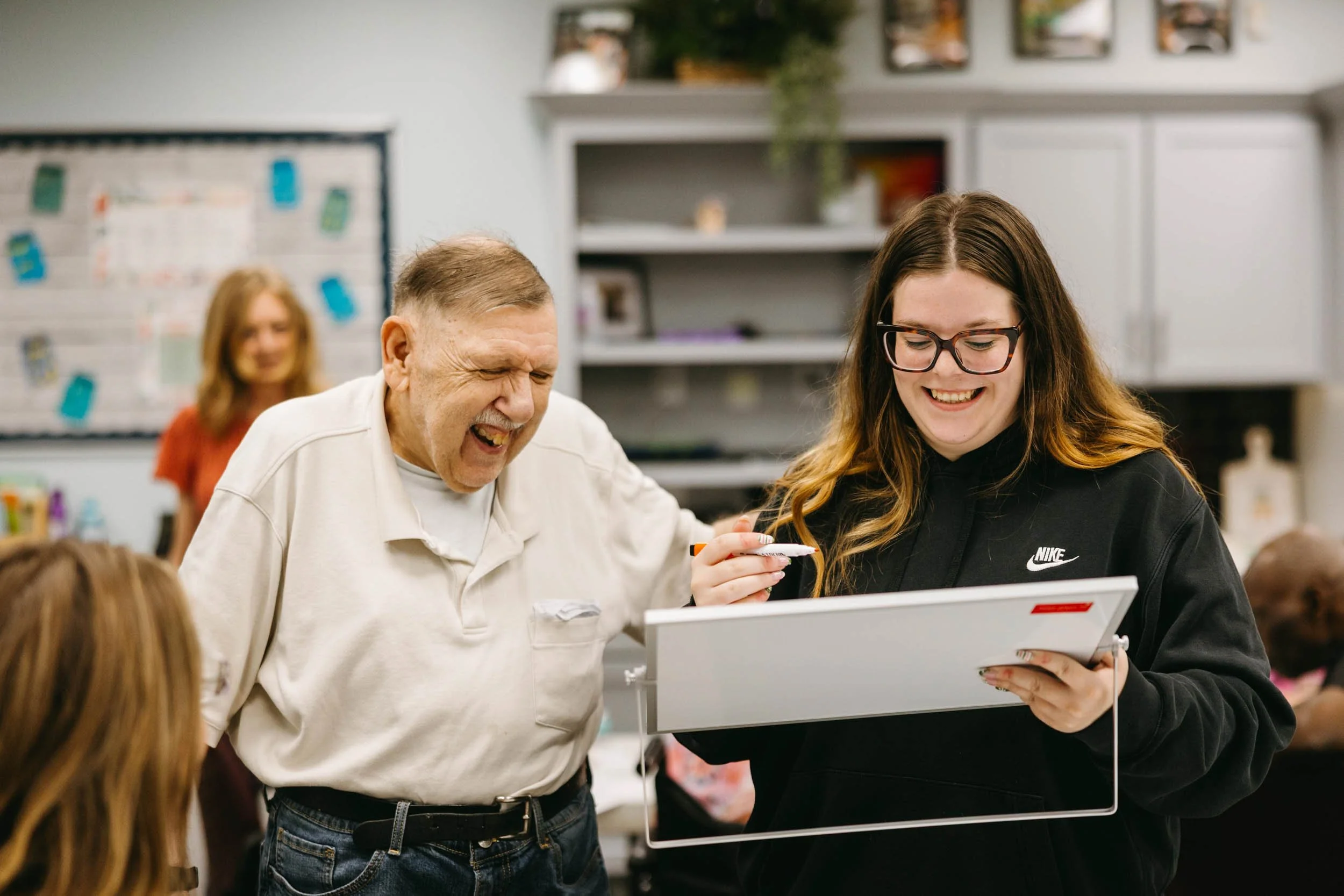 A young woman and an older man are smiling and laughing while looking at a tablet in a classroom. The woman is wearing glasses and a black Nike hoodie, and the man is wearing a white shirt. A woman with long hair and a red shirt is blurred in the background.