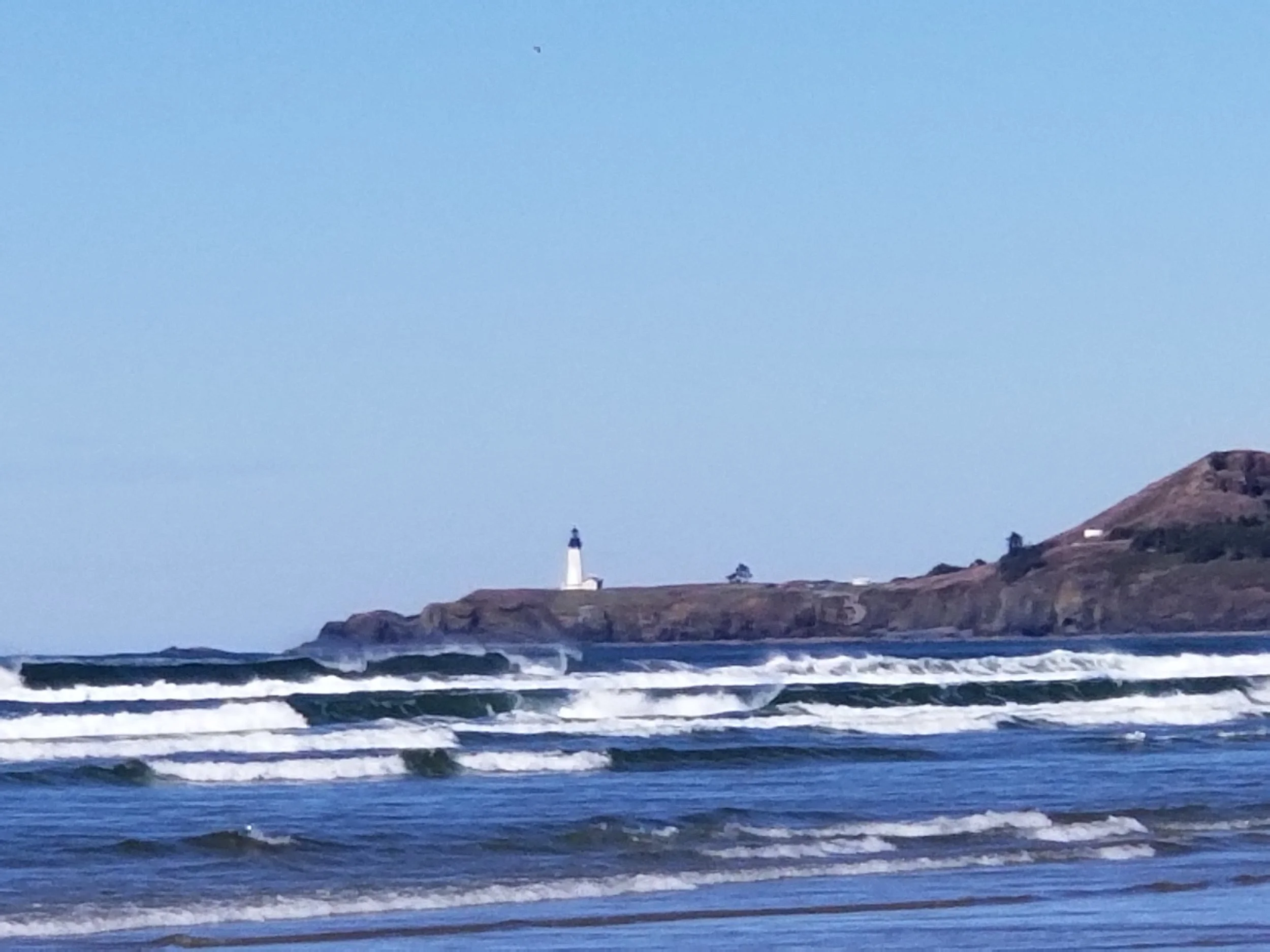 A lighthouse standing on a rocky hill near the ocean, with waves hitting the shore, and a clear blue sky in the background.