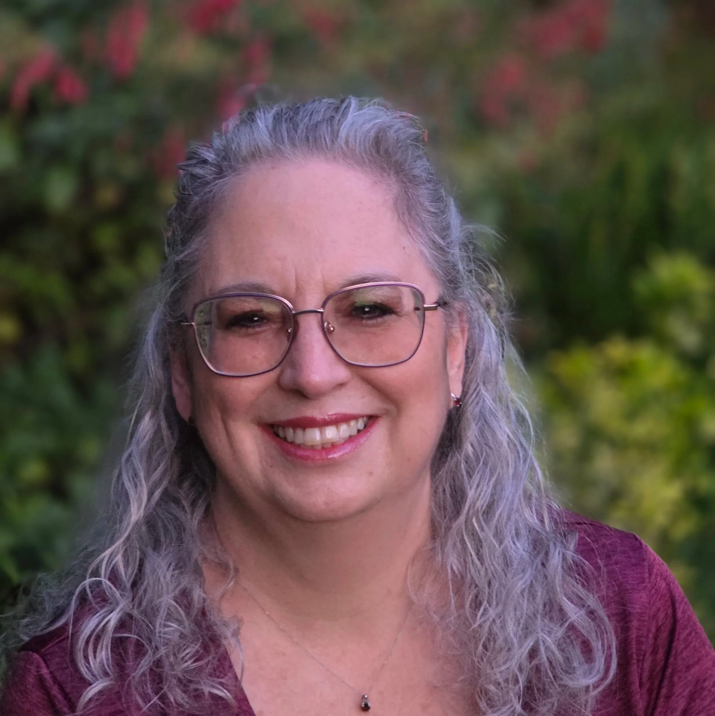 A smiling woman with gray curly hair and glasses, wearing a maroon sweater and a belly button piercing, standing outdoors with blurred green and red foliage in the background.