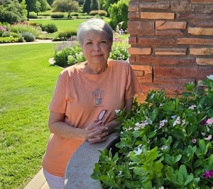 An elderly woman with short gray hair standing outside near a stone wall and green bushes with pink flowers, holding glasses in her hands.