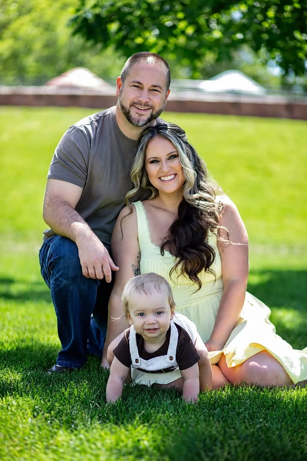 A family of three posing outdoors on green grass, with trees and a fence in the background. The man is kneeling on the left, smiling, with his arm around the woman. The woman, sitting on the grass, has long wavy hair and is smiling at the camera. The toddler girl is in front, crawling on the grass, also smiling.