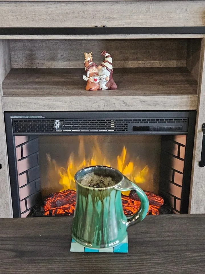 A mug of coffee on a coaster in front of an electric fireplace with artificial flames. There are holiday figurines on a shelf above the fireplace.